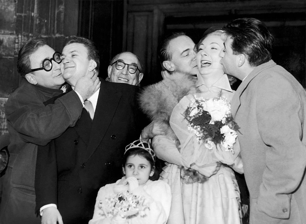 Le réalisateur Marcel Achard embrasse le marié Pierre Mondy tandis que les acteurs René Simon et François Perier embrassent la mariée Claude Gensac lorsqu'ils sortent de l'église le 26 octobre 1951 en France. | Photo : Getty Images