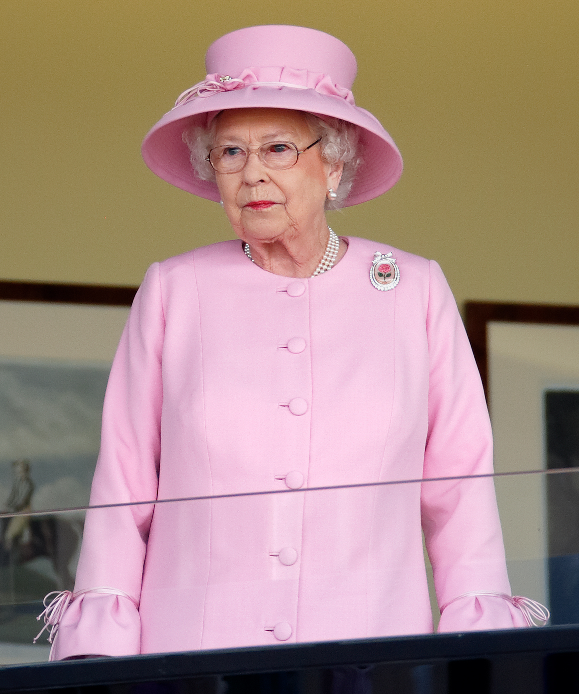 Depuis la loge royale, la reine Elizabeth II assiste aux courses, les yeux rougis, parée de perles et arborant la broche Centenary Rose. Son expression sereine reflète son sens du devoir, malgré un vaisseau sanguin visiblement éclaté lors de cette réunion mondaine.