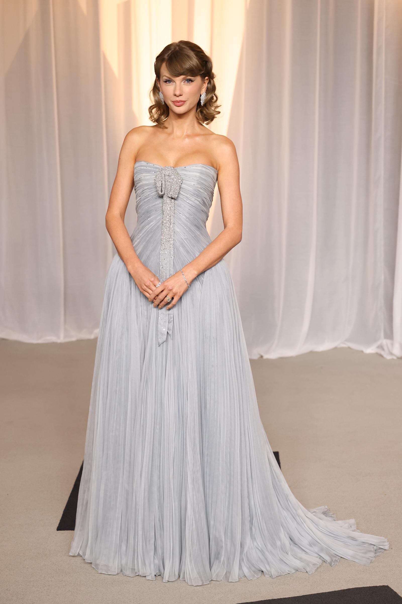 Taylor Swift pose dans une robe plissée bleu glacier sans bretelles, ornée d'un nœud recouvert de cristaux et d'une longue traîne, qu'elle accessoirise avec des boucles d'oreilles étincelantes, un bracelet et des bagues.