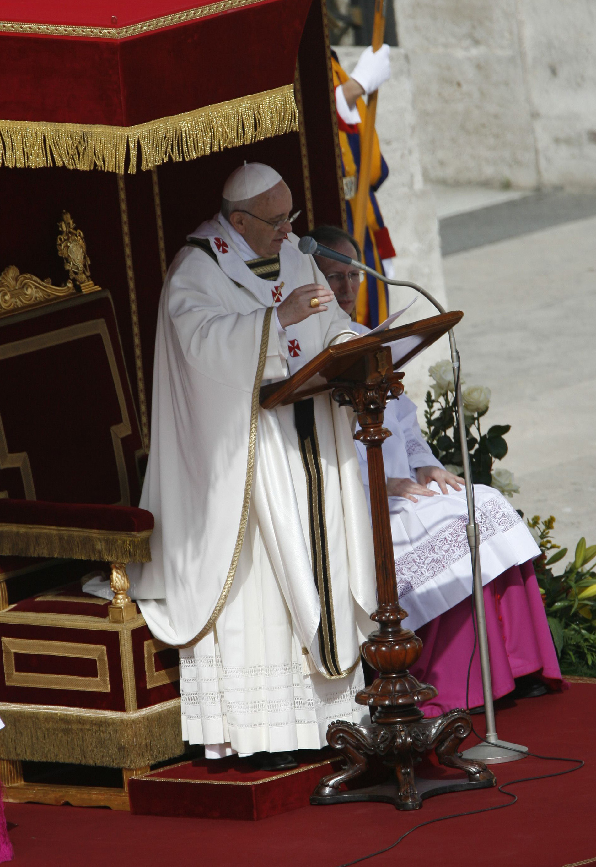 Le pape François prononce un sermon depuis un pupitre en bois sculpté, vêtu d'habits crème avec des croix rouges, debout à côté d'une chaise en forme de trône garnie d'or.