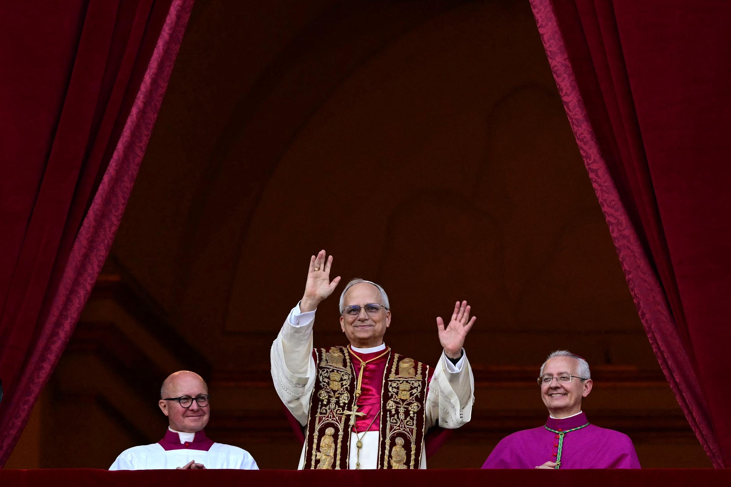 Le pape Léon XIV lève les deux mains en signe de salutation. Il porte une étole richement brodée sur une robe crème et s'avance sur le balcon encadré par des rideaux de velours rouge foncé.