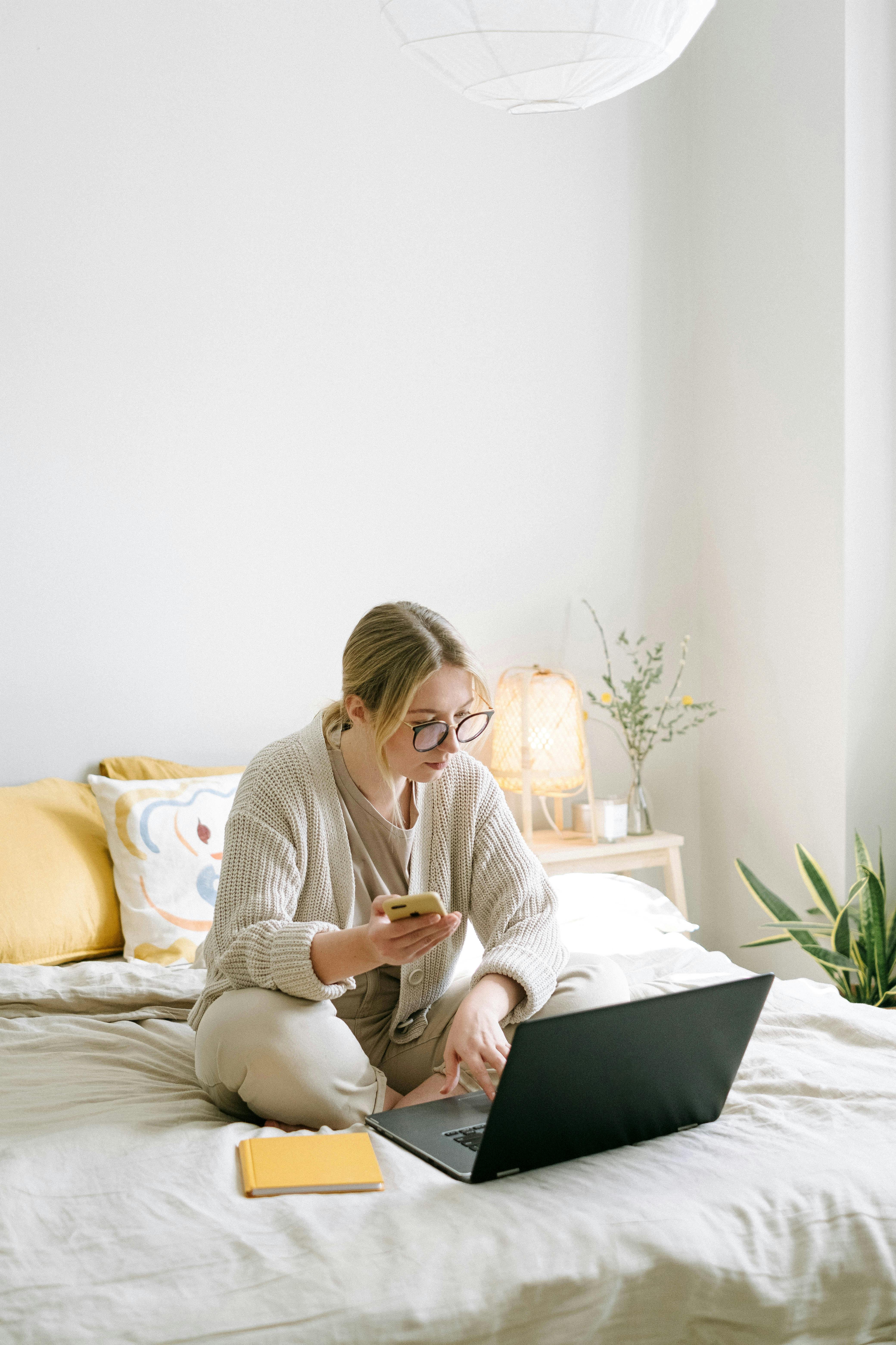 Une femme assise sur le lit avec son téléphone et son ordinateur portable | Source : Pexels