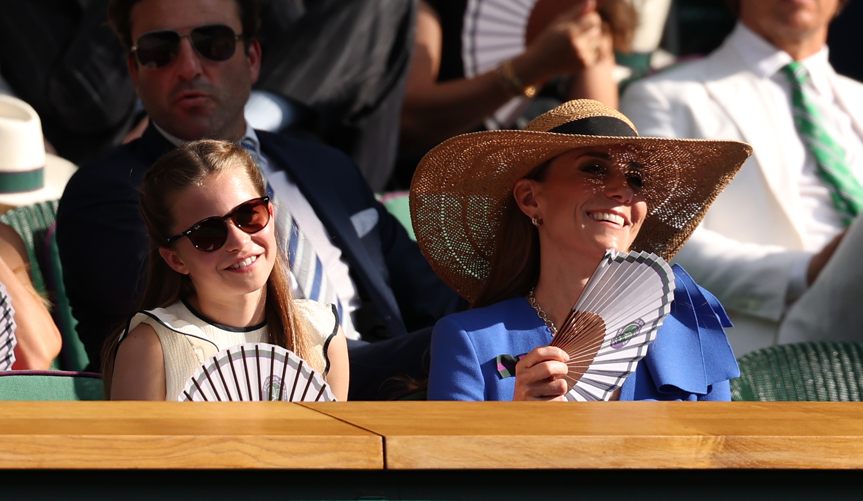 La princesse Charlotte de Galles et Catherine, princesse de Galles, sourient tout en se rafraîchissant avec des éventails en papier dans la loge royale, lors de la quatorzième journée du tournoi de Wimbledon, à l'All England Lawn Tennis and Croquet Club, le 13 juillet 2025, à Londres, en Angleterre. Charlotte regarde la finale du simple messieurs avec des lunettes de soleil en écaille foncée et un haut sans manches crème à volants, assise à côté de Catherine, qui arbore un look estival avec une robe bleu vif, des lunettes de soleil tendance et un chapeau de paille à large bord. | Source : Getty Images