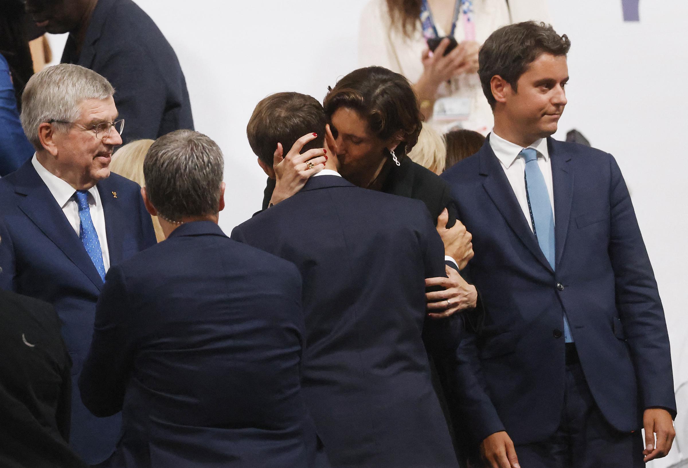 Amélie Oudéa-Castéra enlace Emmanuel Macron de ses deux bras, posant une main sur l'arrière de sa tête tout en le serrant de près. Son visage effleure son cou, tandis que plusieurs personnes, dont Thomas Bach, se tiennent en vue.