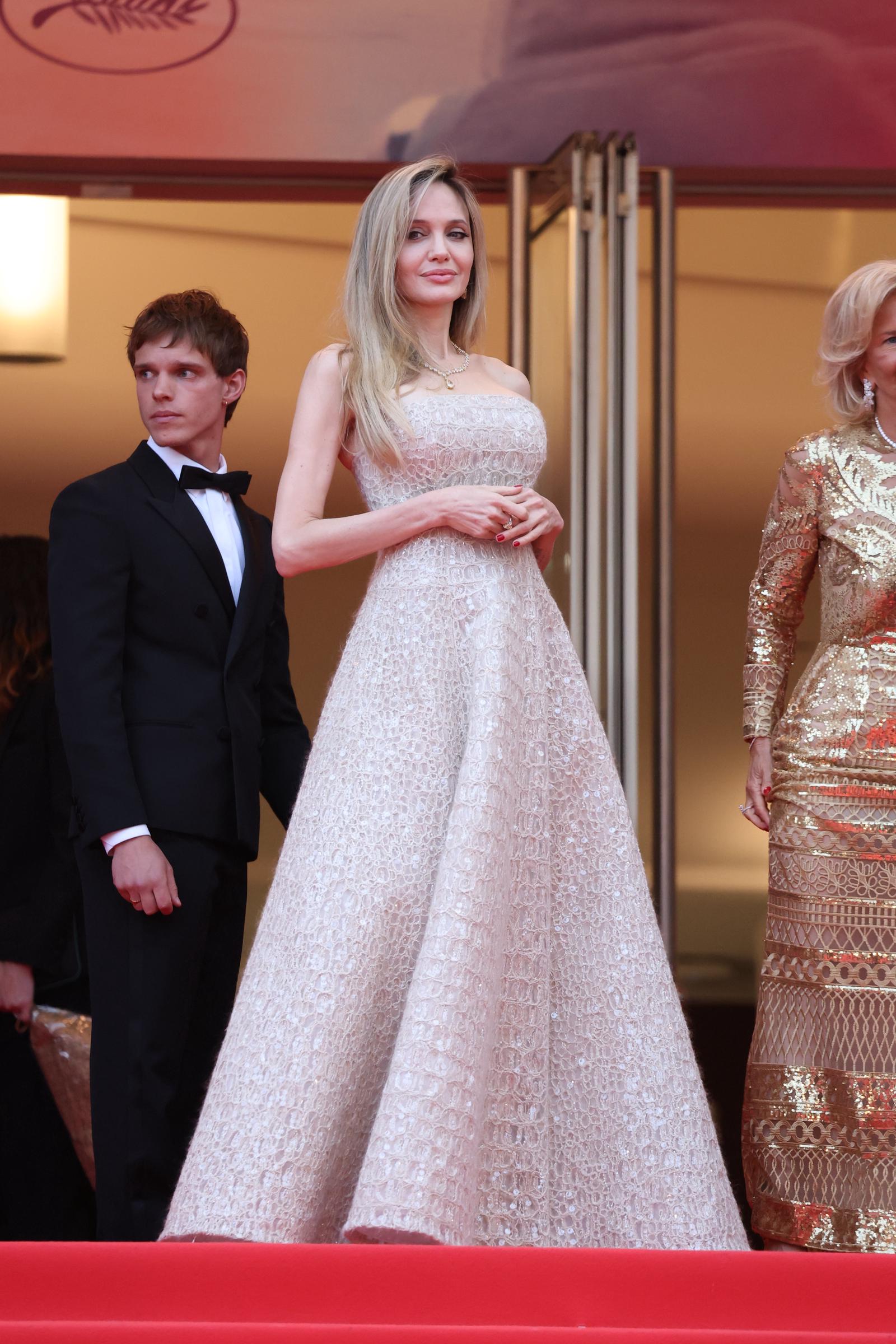 Angelina Jolie se tient debout en haut de l'escalier du Palais des Festivals, les mains délicatement jointes devant sa robe Brunello Cucinelli aux détails complexes, dégageant une assurance intemporelle sur fond de rouge et d'or.