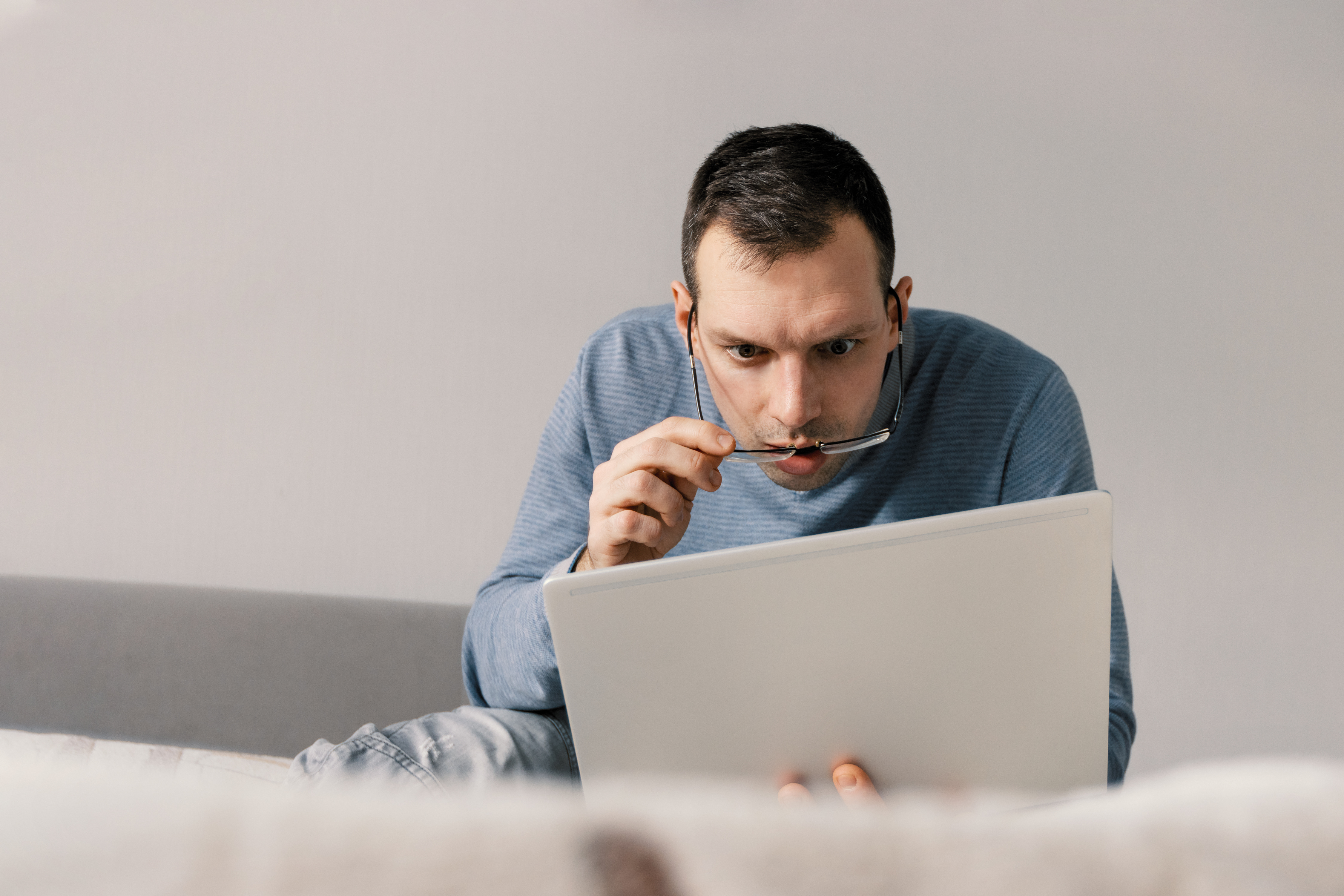 Un homme choqué portant des lunettes et regardant l'écran de son ordinateur portable alors qu'il est assis sur le canapé. Expression faciale - wow | Source : Getty Images