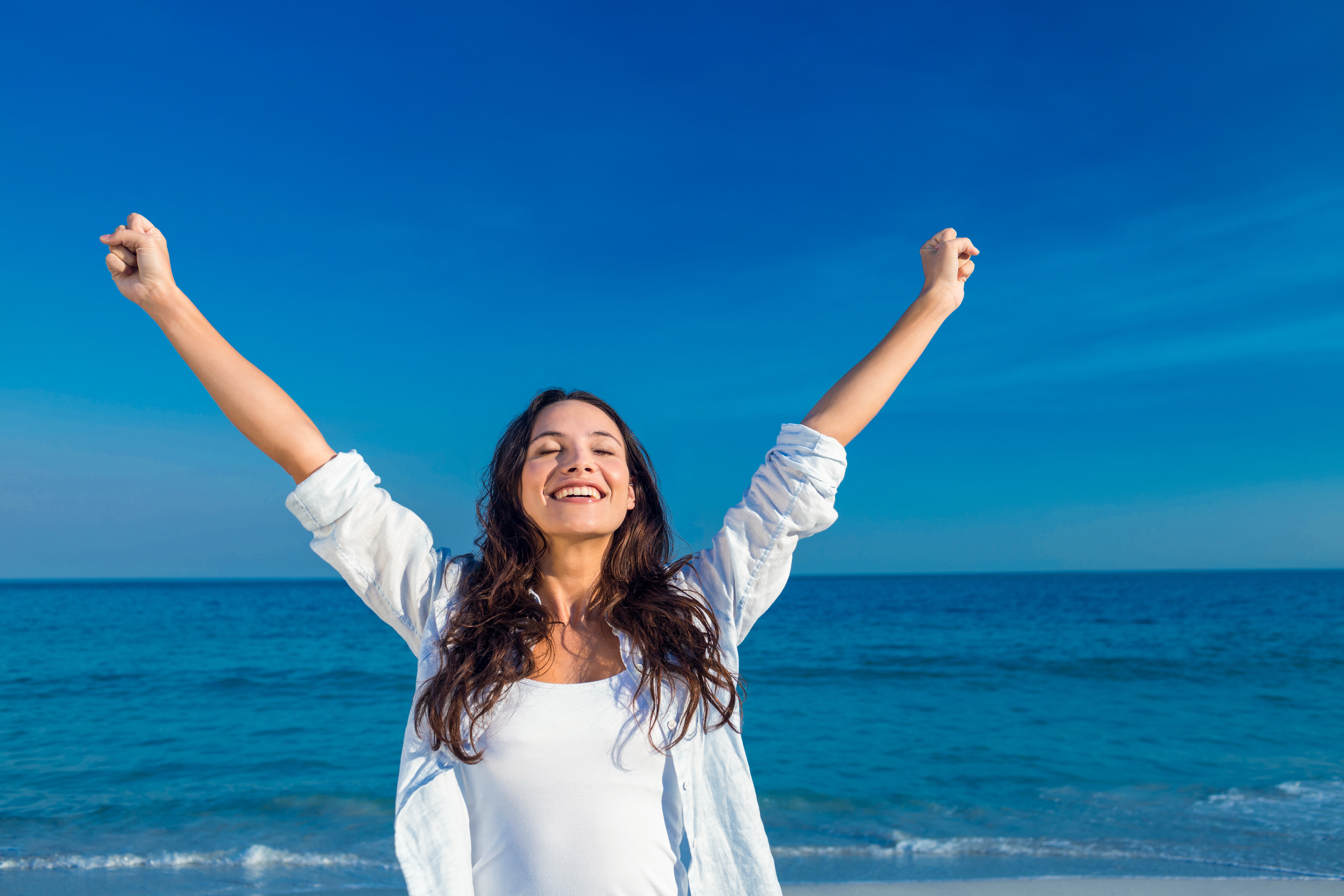 Une femme souriant avec ses mains levées au-dessus de sa tête sur la plage | Source : Shutterstock