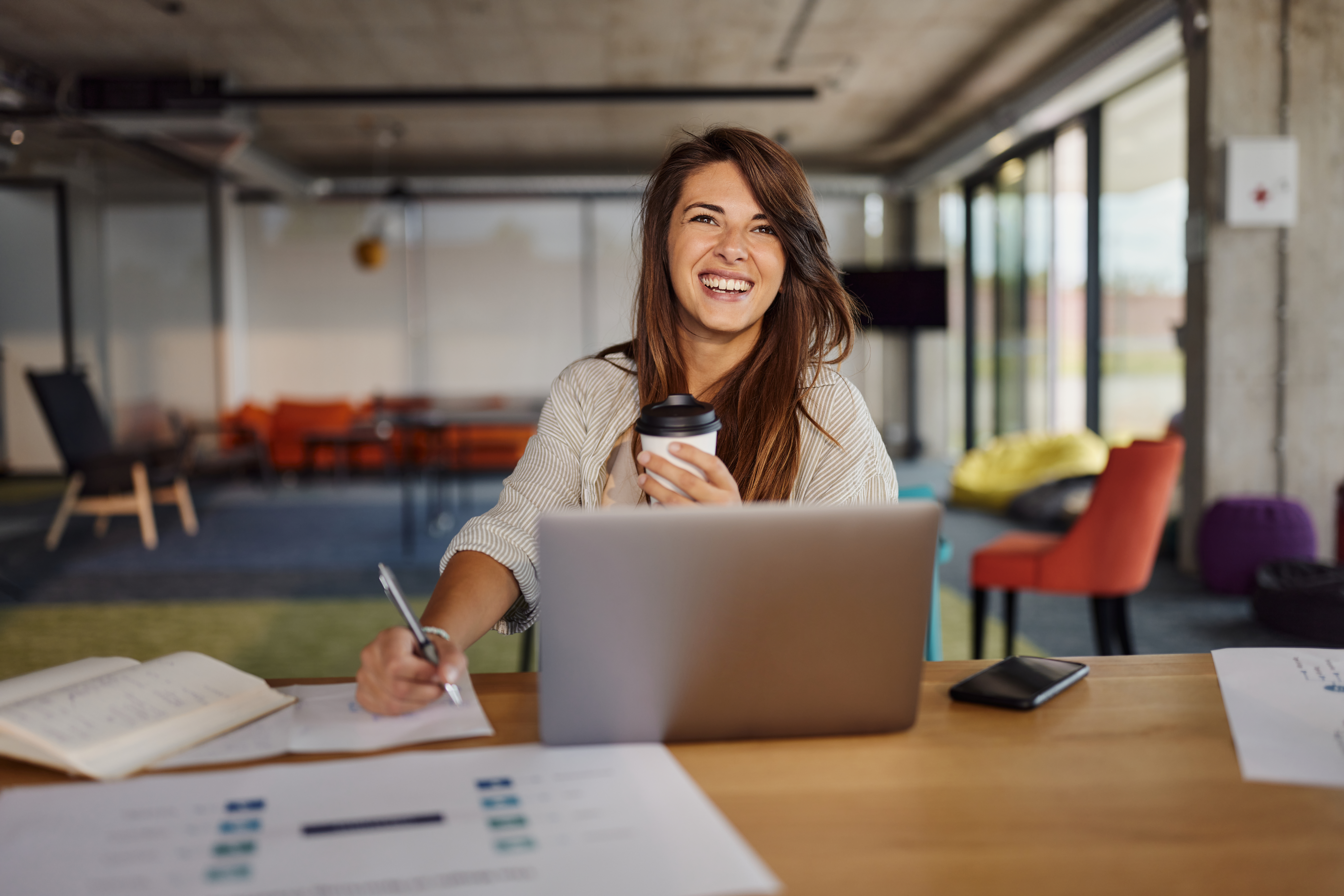 Femme créative heureuse prenant des notes tout en travaillant sur son ordinateur portable au bureau | Source : Getty Images