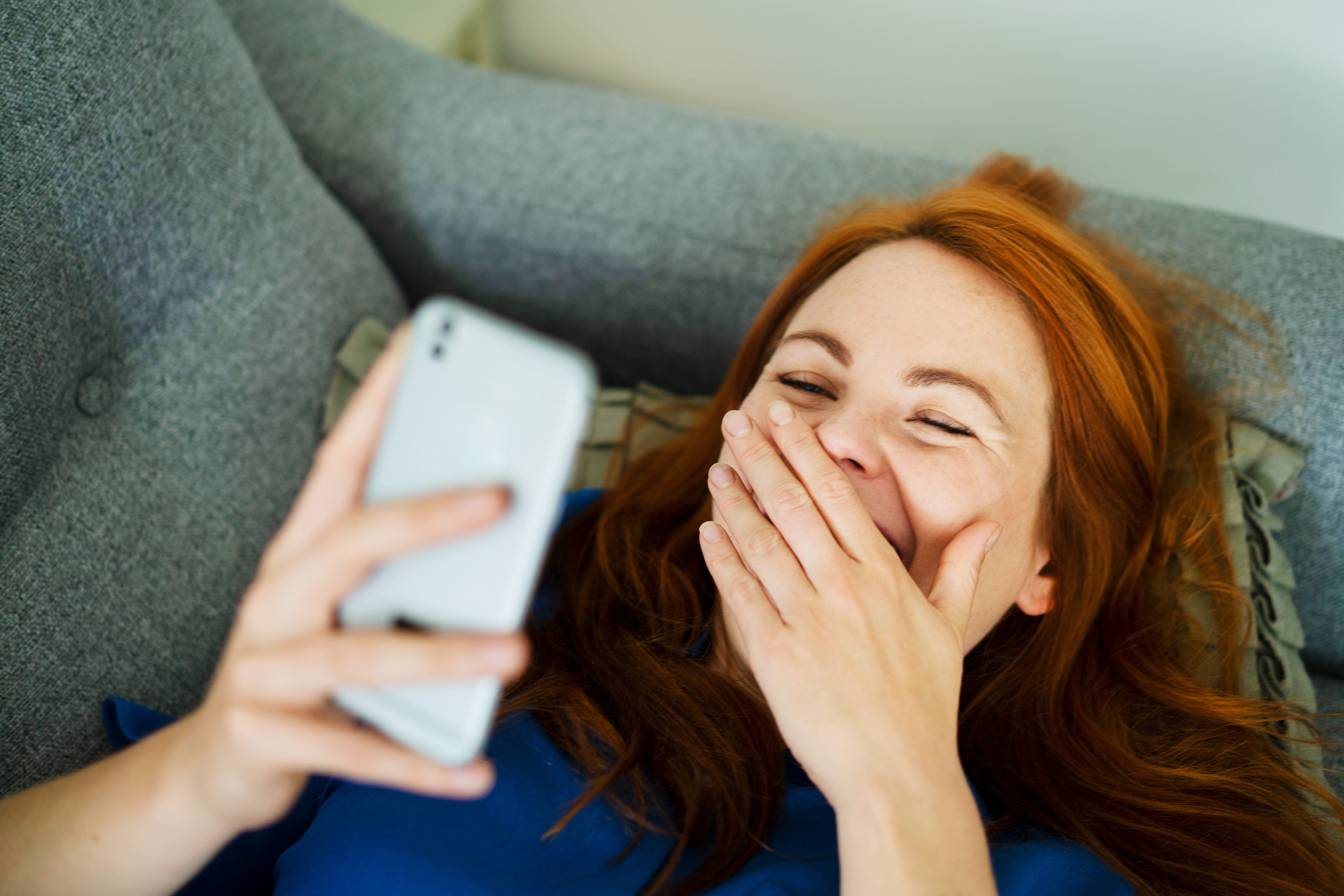 Une femme qui rit devant son téléphone alors qu'elle est allongée sur le canapé | Source : Getty Images