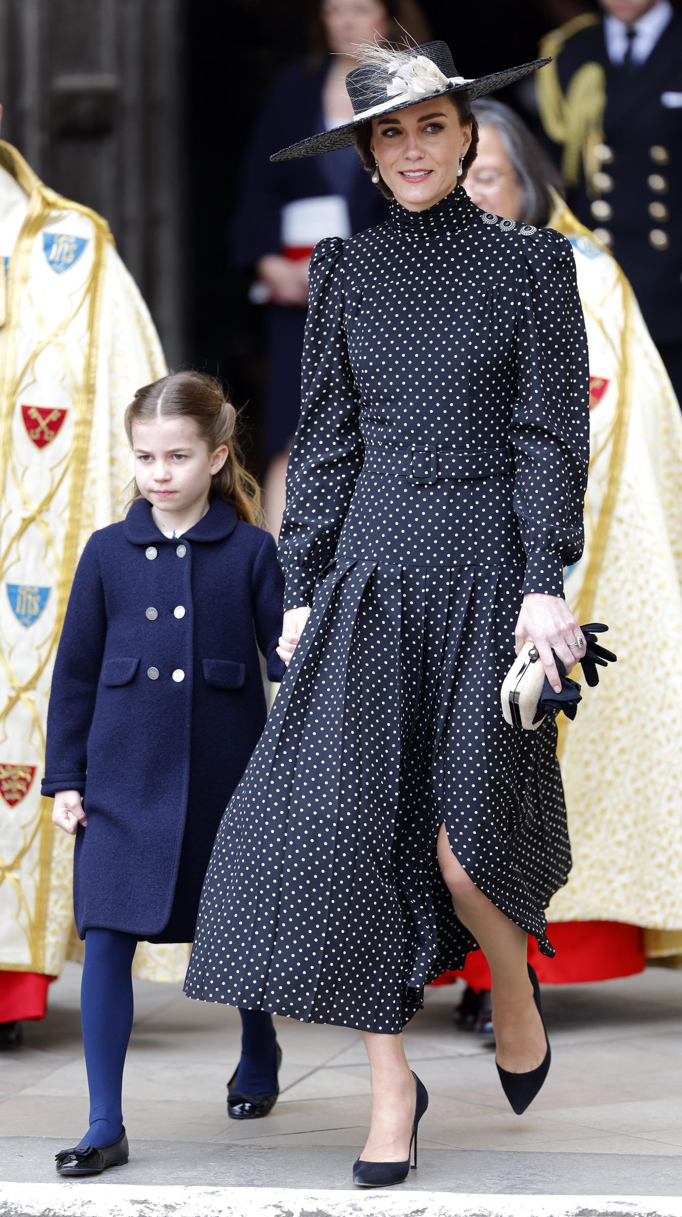 La princesse Charlotte marche main dans la main avec Catherine, duchesse de Cambridge, alors qu'elles quittent l'abbaye de Westminster après la cérémonie d'action de grâce en hommage au prince Philip, duc d'Édimbourg, le 29 mars 2022, à Londres, en Angleterre. Charlotte porte un manteau bleu marine boutonné sur des collants assortis et des chaussures noires vernies, et tient la main de Catherine, qui est vêtue d'une élégante robe midi plissée à pois noirs et blancs, d'un chapeau à large bord orné de motifs floraux et de talons noirs pointus. | Source : Getty Images