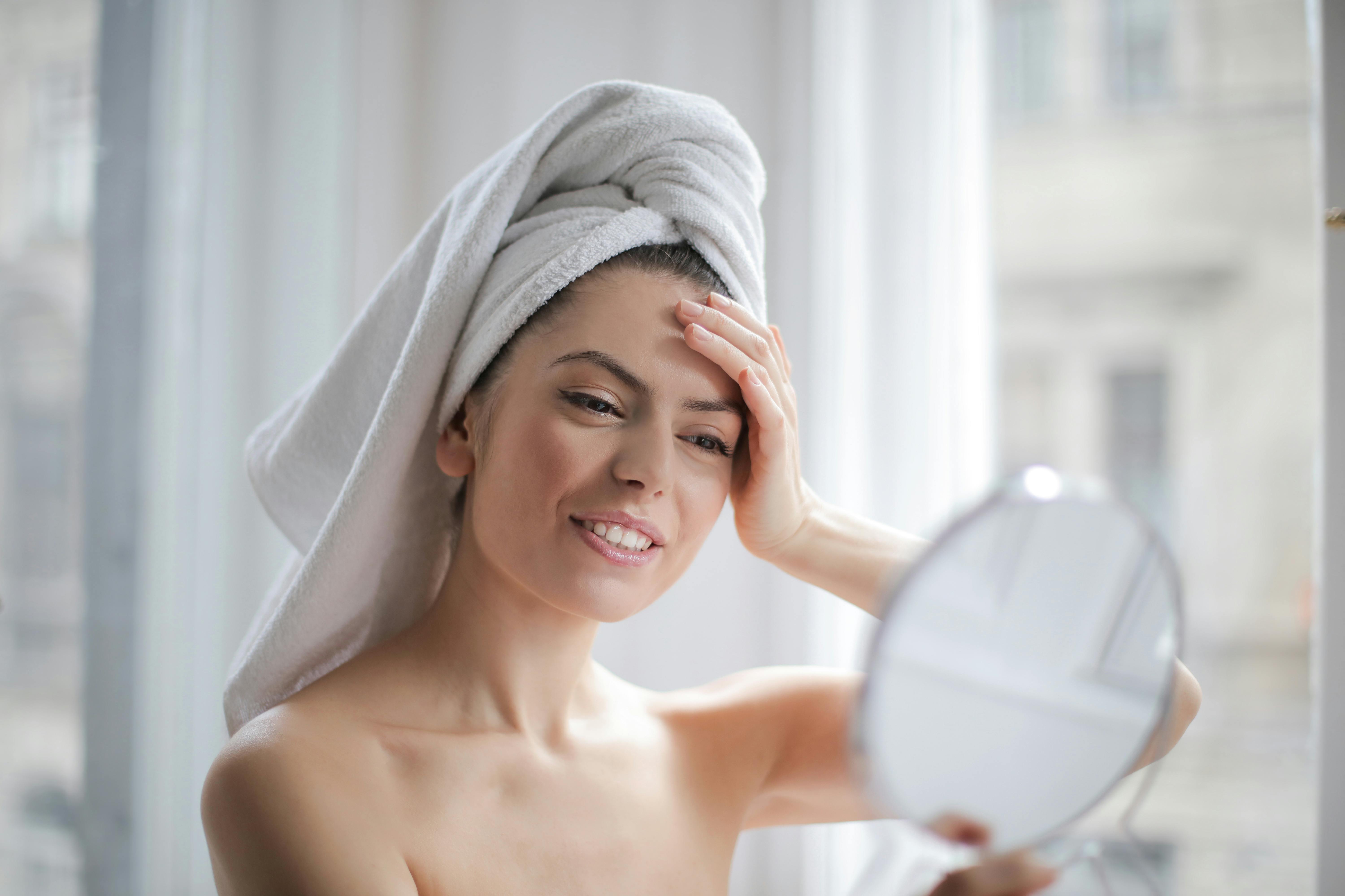 Une femme heureuse se regardant dans le miroir tout en portant une serviette pour se sécher les cheveux | Source : Pexels