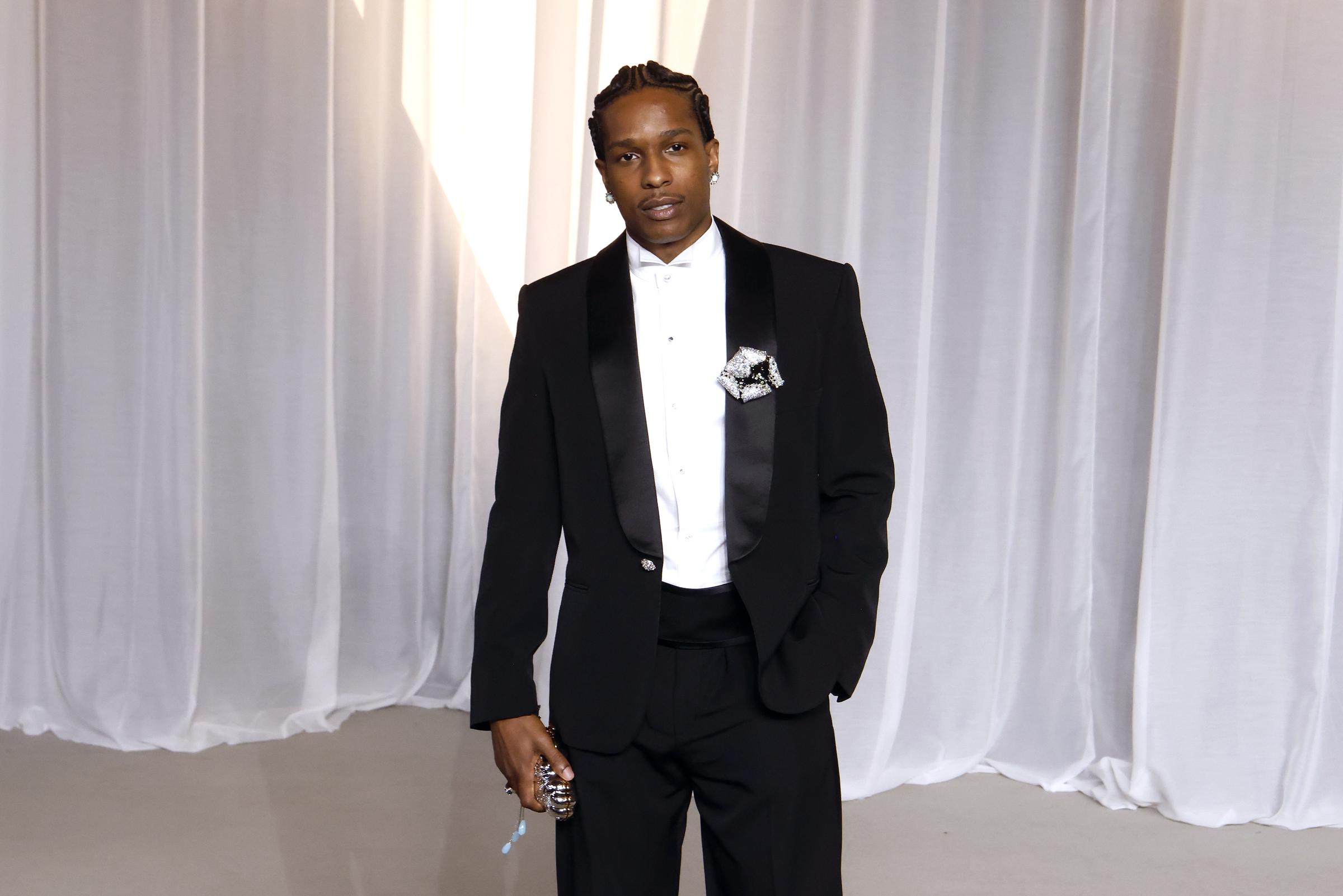 A$AP Rocky incarne le chic moderne en tenue de soirée avec un smoking épuré, taillé sur mesure, doté de larges revers châle en satin et d'une ceinture de smoking haute. Laissant de côté le nœud papillon traditionnel au profit d’une chemise blanche minimaliste à col ailé, le rappeur rehausse cette tenue classique avec une broche florale ouvragée épinglée à son revers, des tresses africaines soignées et une pochette de soirée originale et scintillante.