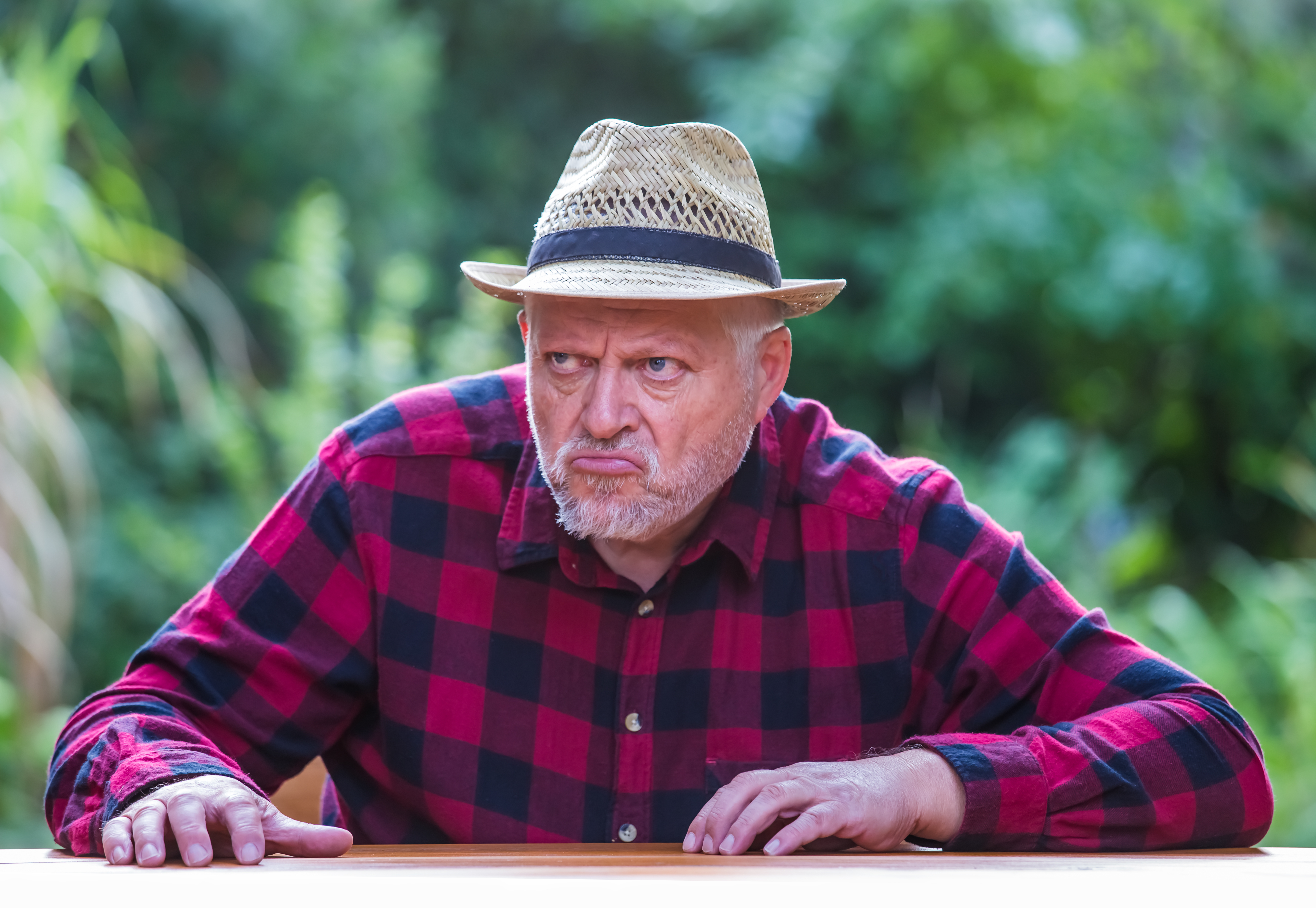 Un homme âgé coiffé d'un chapeau est assis avec mauvaise humeur à une table dans le jardin. | Source : Shutterstock