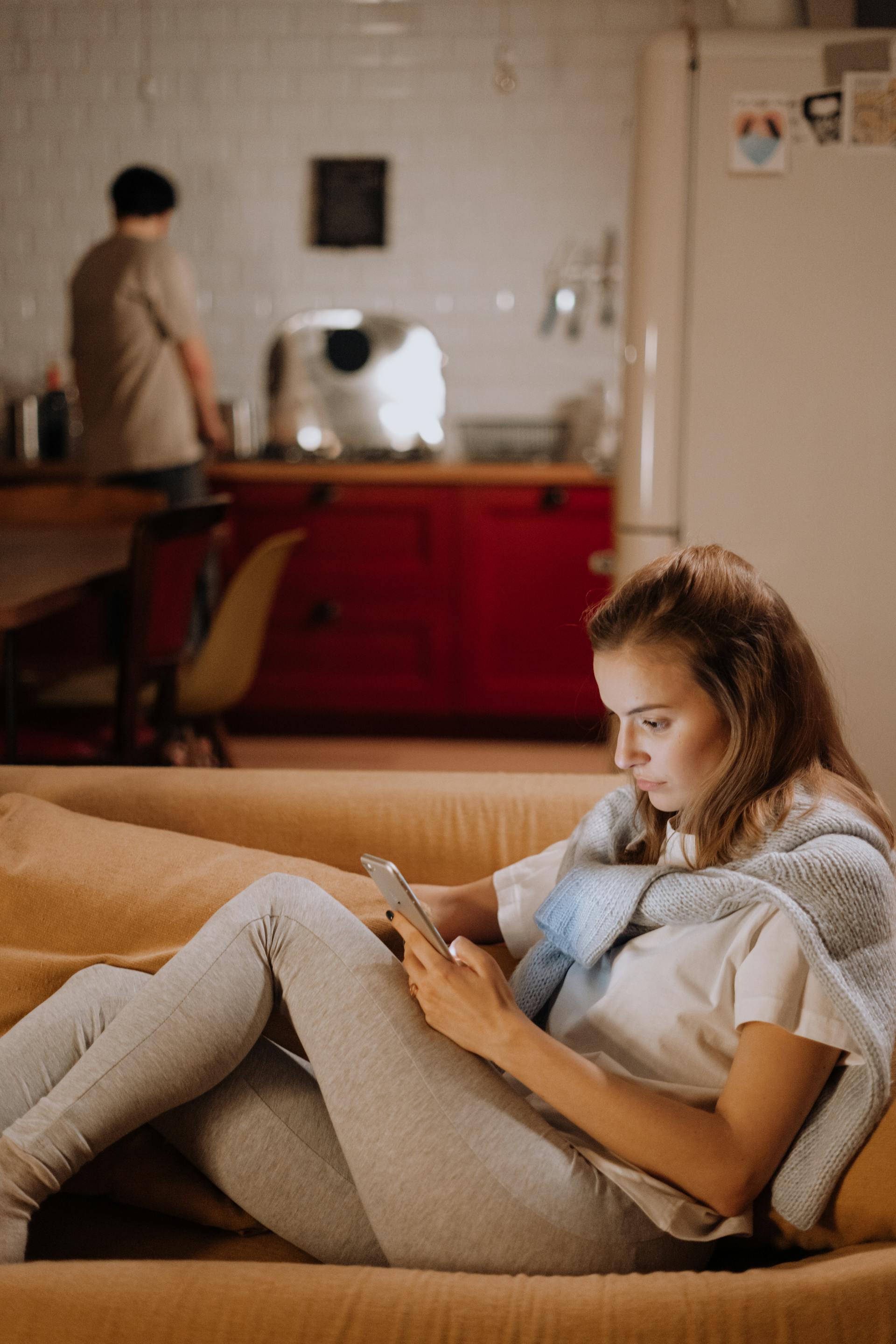 Une femme assise sur le canapé et utilisant son téléphone tandis qu'un homme se tient dans la cuisine | Source : Pexels