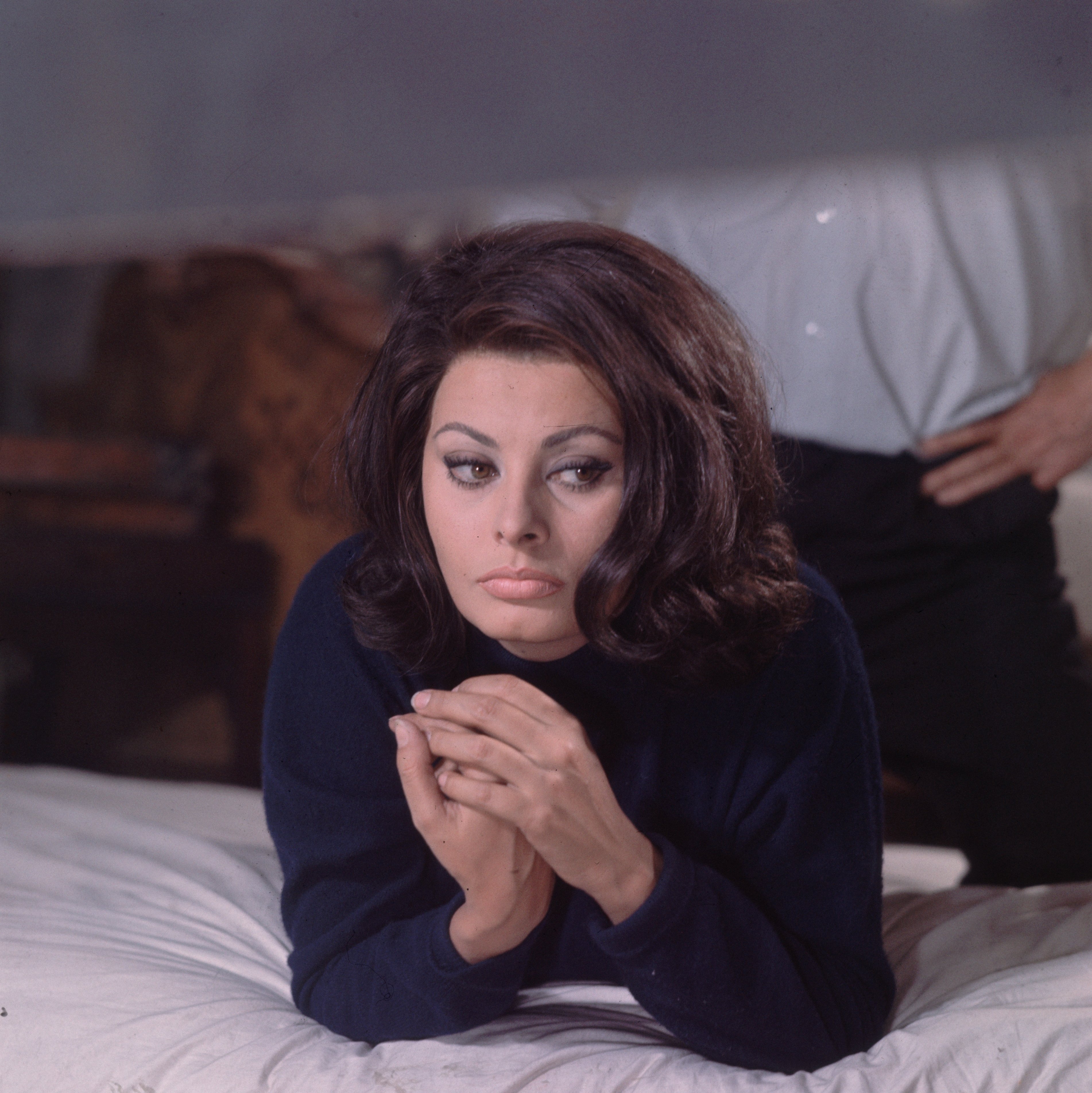 Sophia Loren est allongée sur un lit et porte un pull à col roulé bleu tandis qu'un homme non identifié se tient derrière elle, en novembre 1964, à Londres, en Angleterre. | Photo : Getty Images