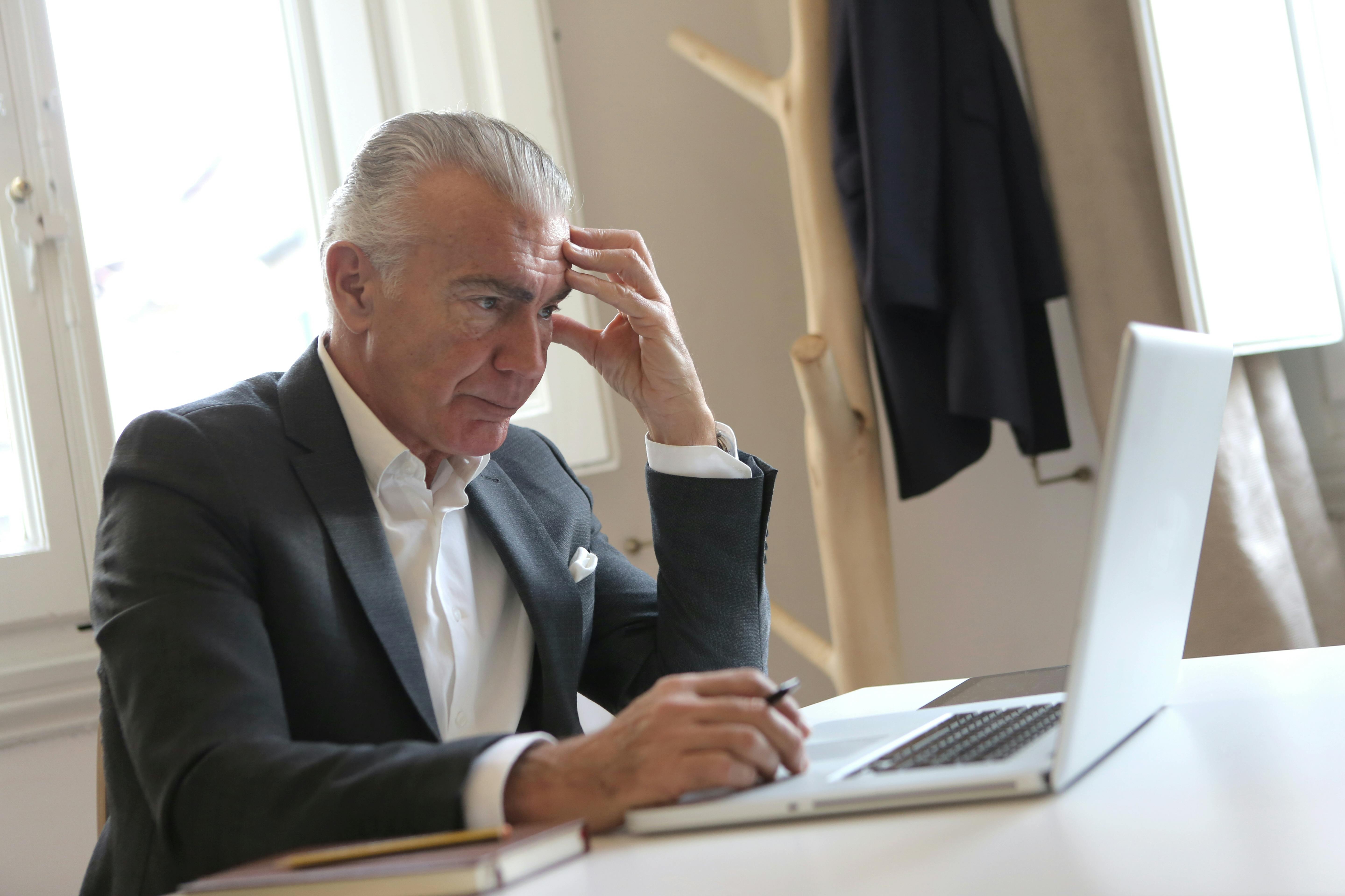 Un homme âgé stressé assis avec sa main sur sa tempe tout en tenant un stylo et en regardant son ordinateur portable | Source : Pexels