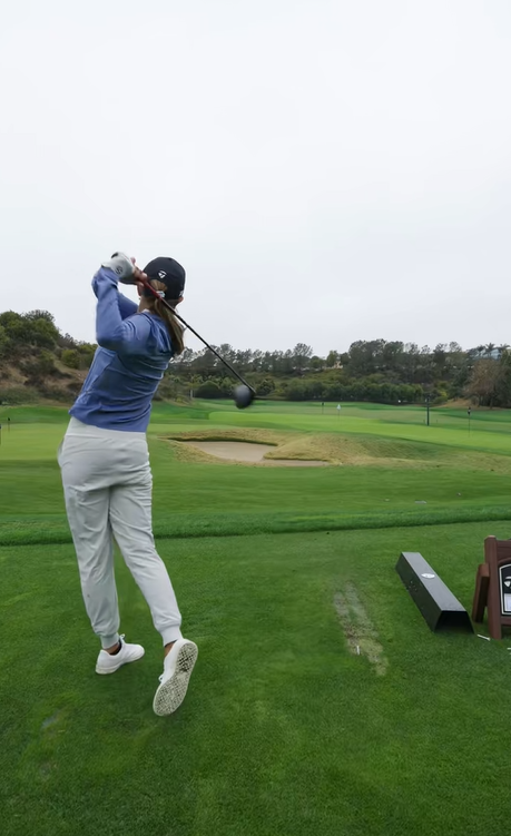 Kai Trump termine son swing sur le terrain de golf, vêtu d'un haut bleu, d'un pantalon blanc et d'une casquette noire par un temps couvert | Source : Tiktok/thekaitrumpgolfer