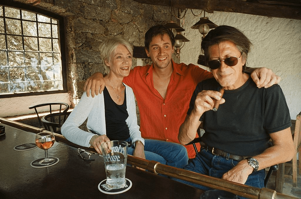Thomas DUTRONC tenant par les épaules ses parents Jacques DUTRONC et Françoise HARDY assis au comptoir du bar installé sur la terrasse de leur maison de MONTICELLO en Haute-Corse. | Photo : Getty Images