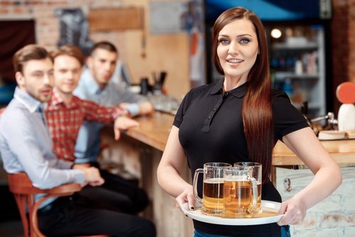Une belle serveuse tenant un plateau avec des verres à bière pendant que les jeunes hommes la regardent avec étonnement | Photo : Shutterstock
