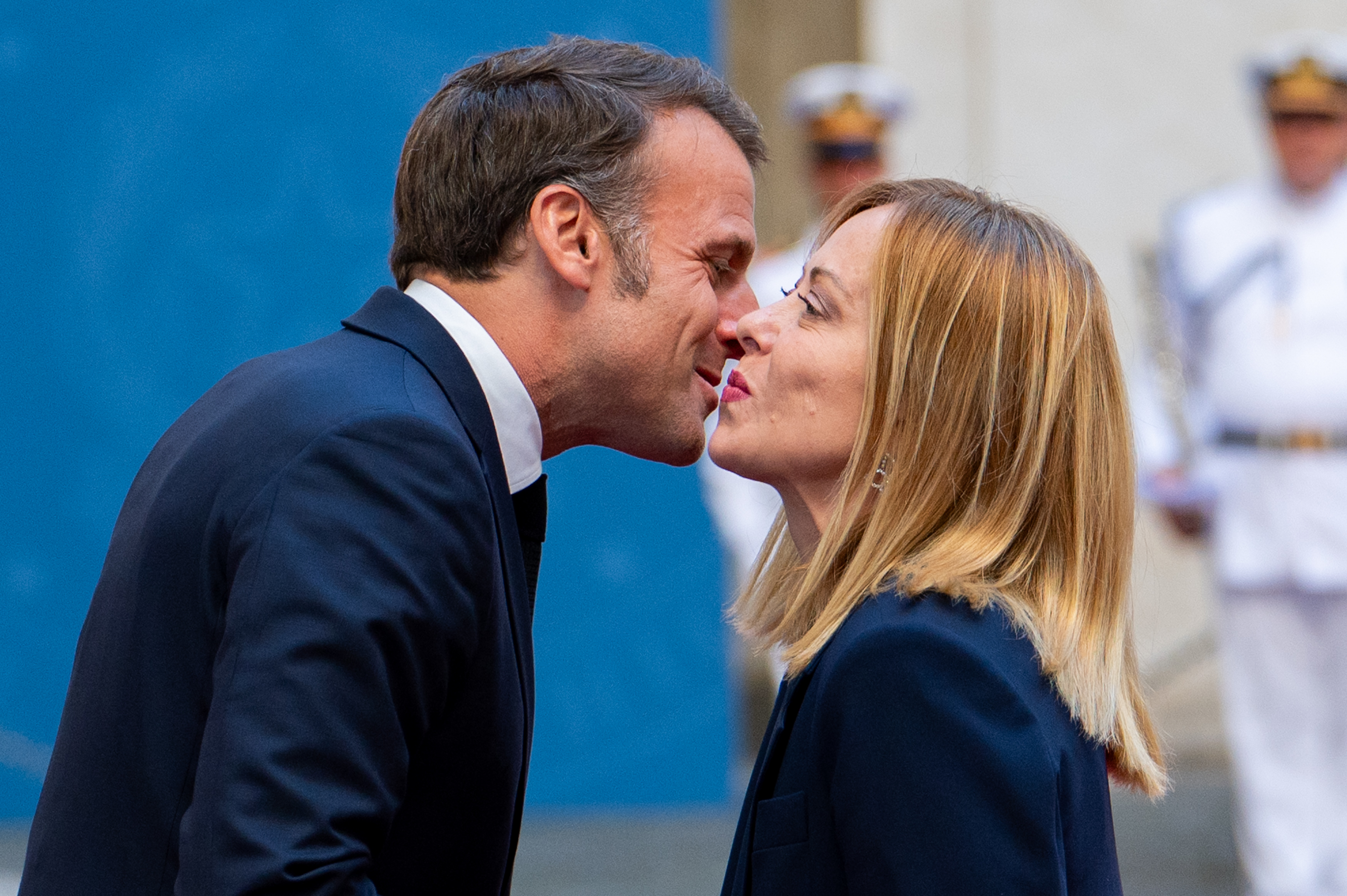 Emmanuel Macron se penche pour saluer Giorgia Meloni avec un baiser traditionnel sur la joue alors qu'ils se tiennent étroitement face à face. Les deux dirigeants maintiennent un contact visuel direct, le personnel en uniforme étant flou à l'arrière-plan.