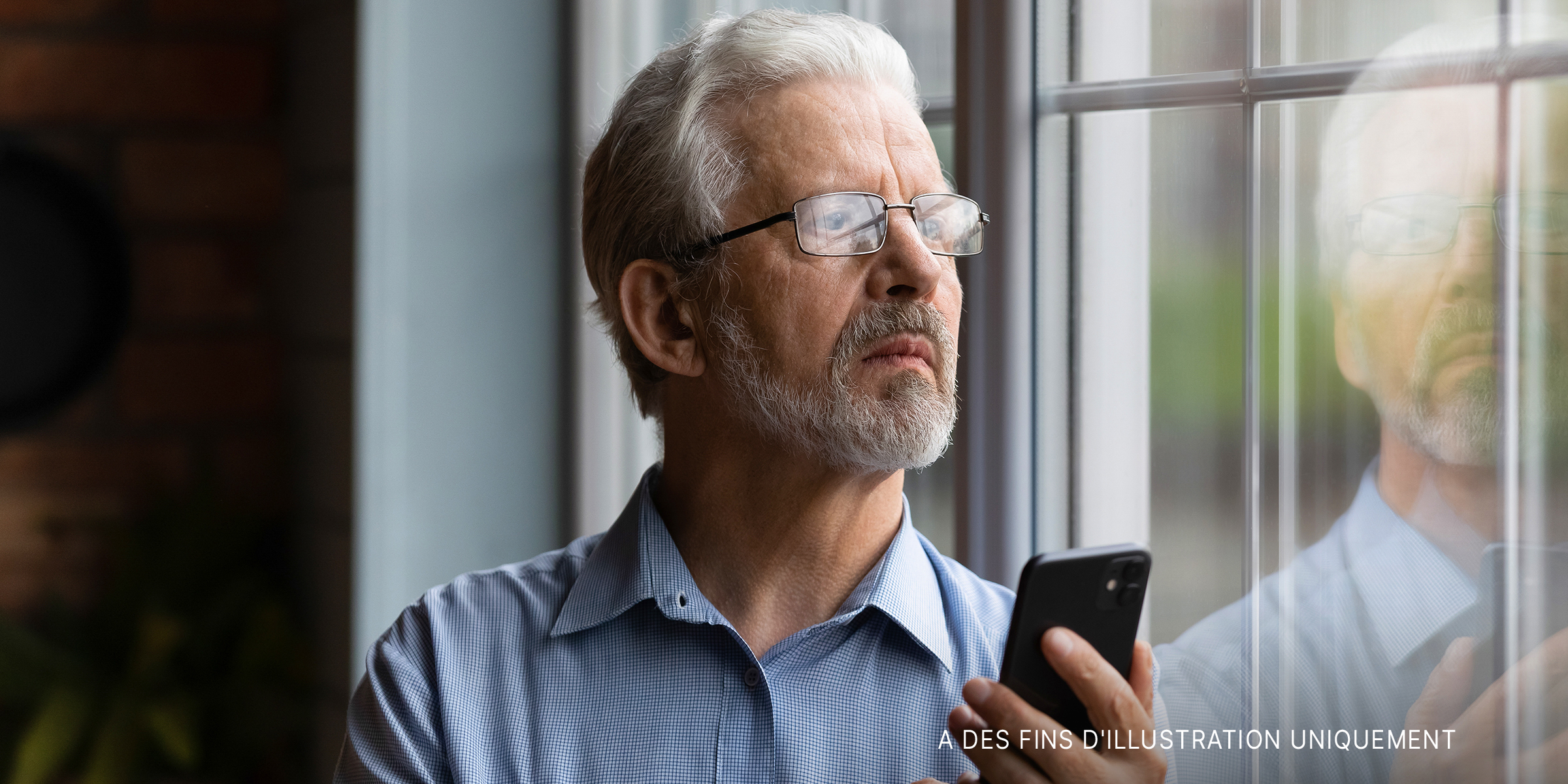 Un homme d'un certain âge tient un téléphone tout en restant debout et en regardant à l'extérieur d'une fenêtre | Source : Shutterstock