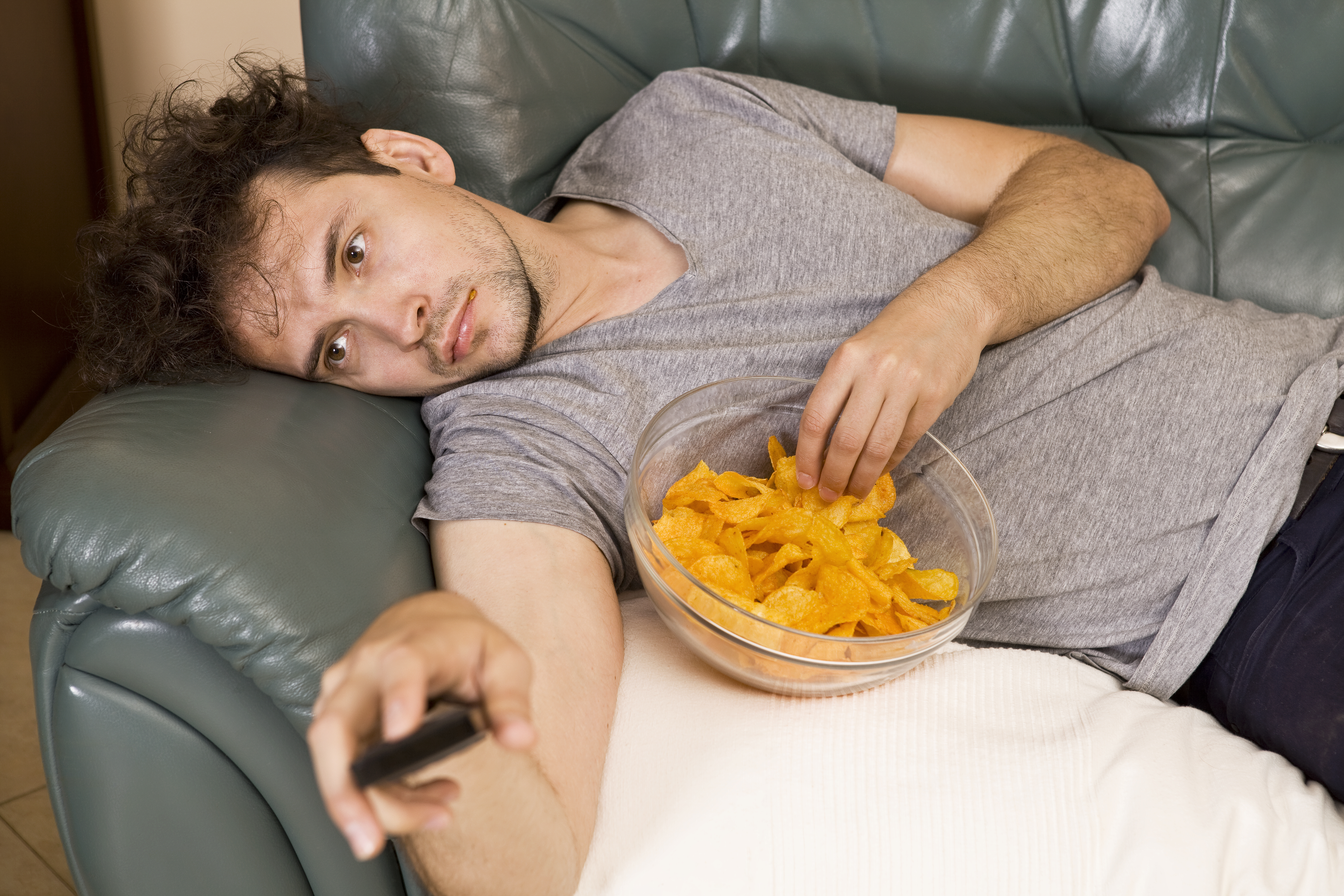 Un homme paresseux allongé sur un canapé regardant la télévision tout en mangeant des snacks | Source : Getty Images