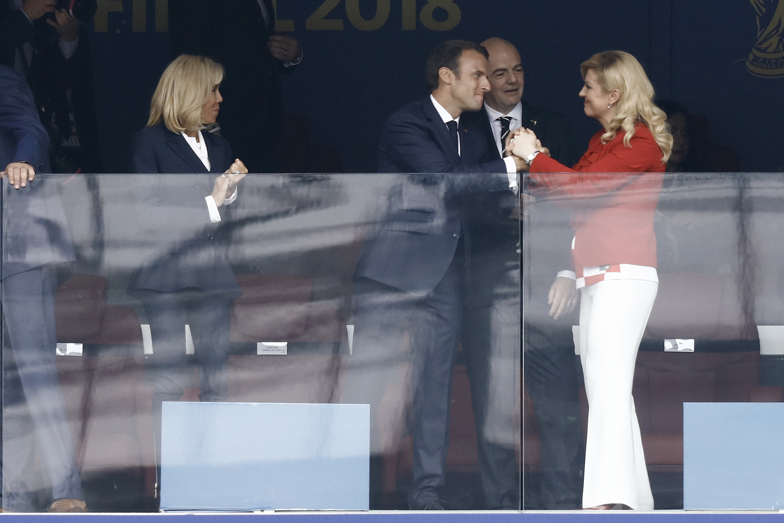 Emmanuel Macron est vu en train de saluer chaleureusement Kolinda Grabar-Kitarović avec une poignée de main ferme tout en souriant, tandis que Brigitte Macron regarde sur le côté. L'interaction a lieu dans une loge d'observation pendant la finale de la Coupe du monde de football 2018.