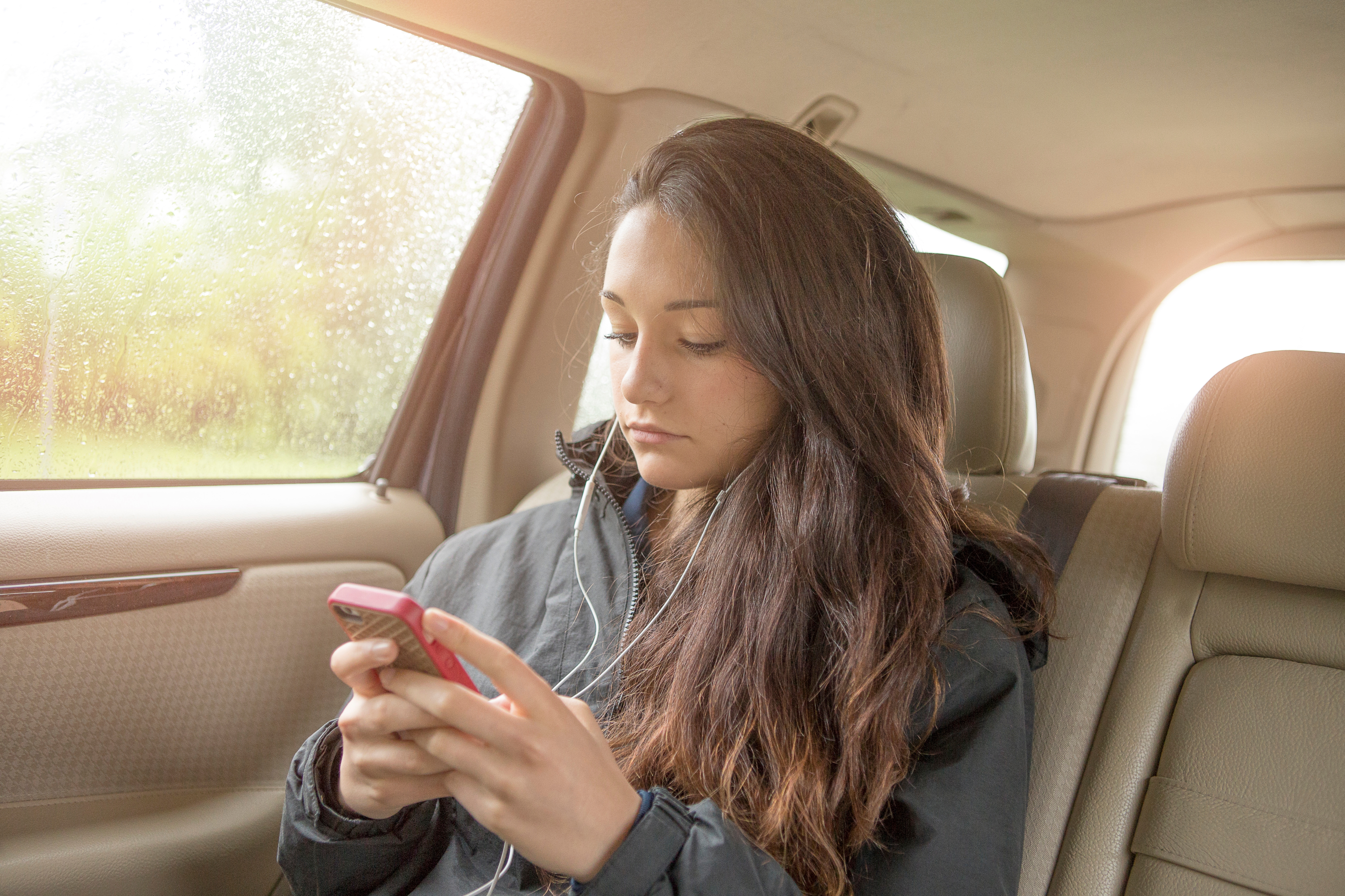 Adolescente maussade sélectionnant la musique de son smartphone sur le siège arrière d'une voiture | Sources : Getty Images