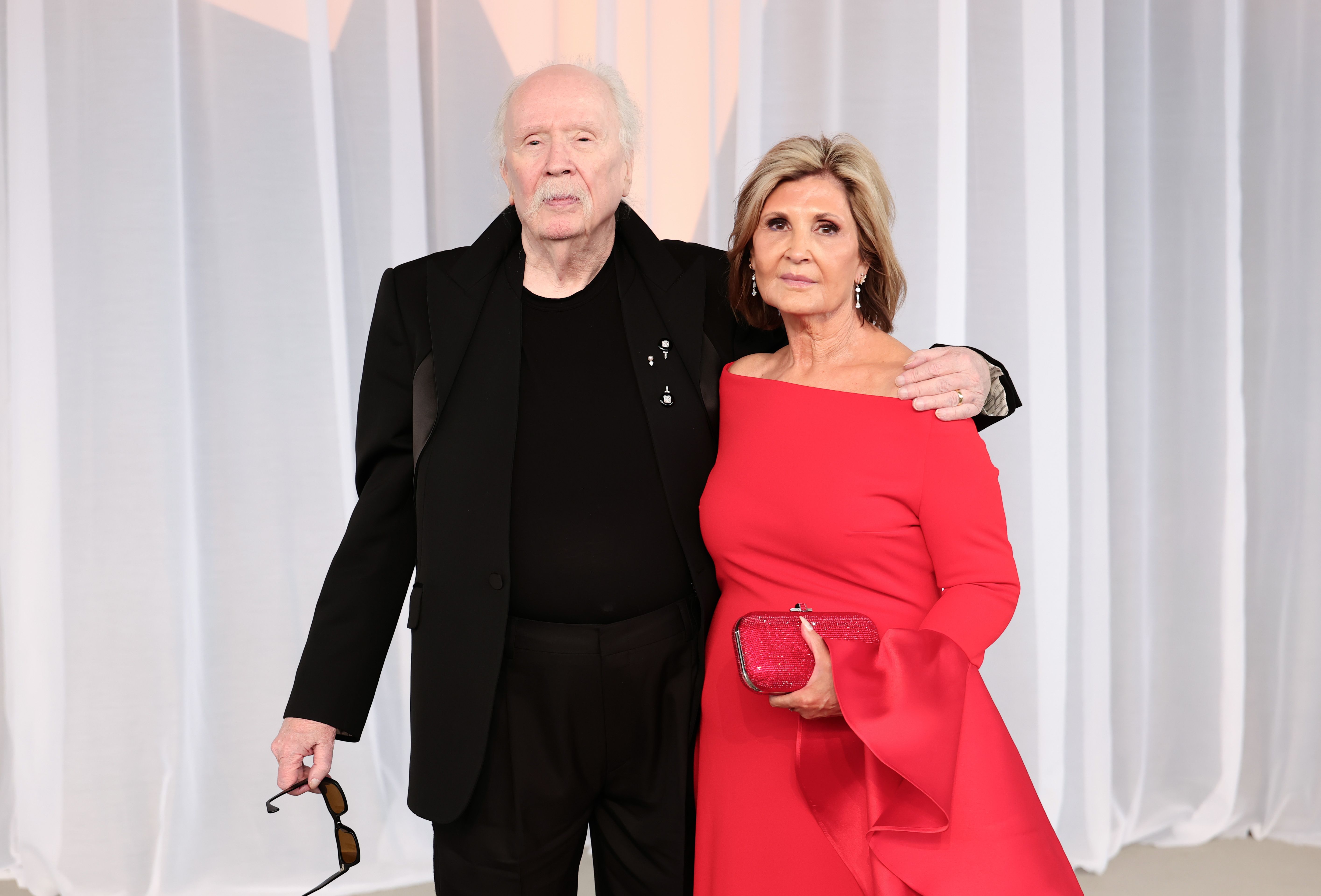 John Carpenter et sa femme, la productrice Sandy King, posent ensemble dans des tenues assorties. Sandy King se démarque dans une robe rouge cerise éclatante aux épaules dénudées, dotée de manches cloche spectaculaires et assortie d'une pochette rubis scintillante, tandis que John Carpenter opte pour un costume sur mesure entièrement noir aux revers bien dessinés et un pull à col rond foncé assorti, tenant ses lunettes de soleil à la main alors qu'il célèbre la remise du Luminary Award.