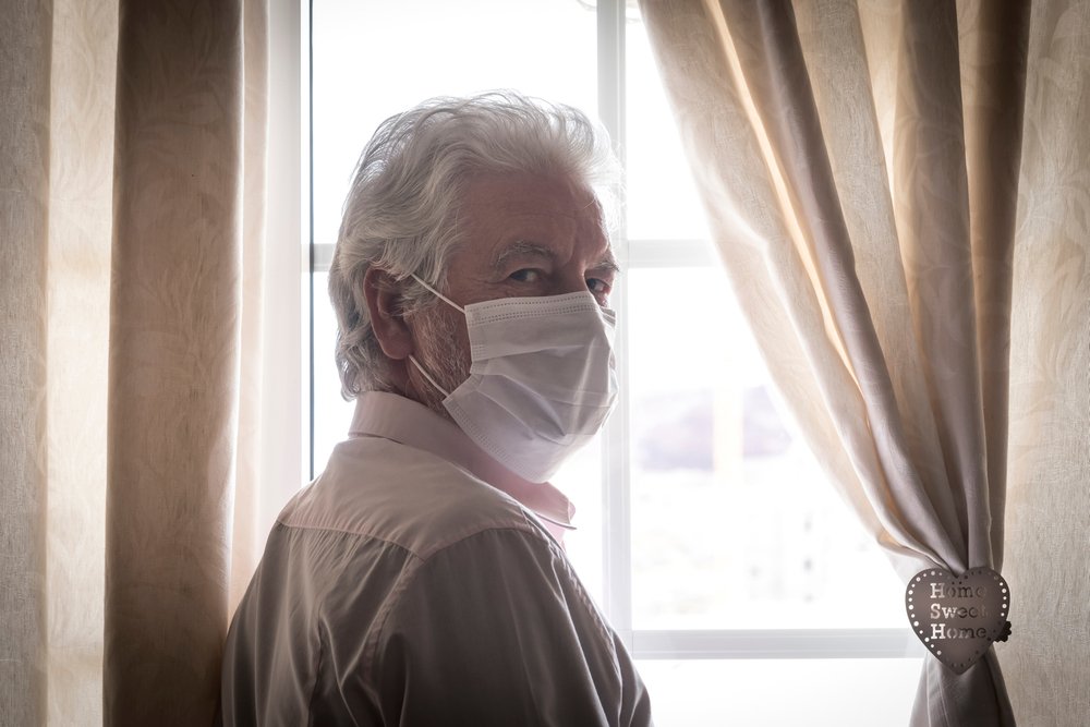  Un homme âgé portant un masque de protection derrière la fenêtre reste à la maison pour éviter la contagion | Source : Shutterstock