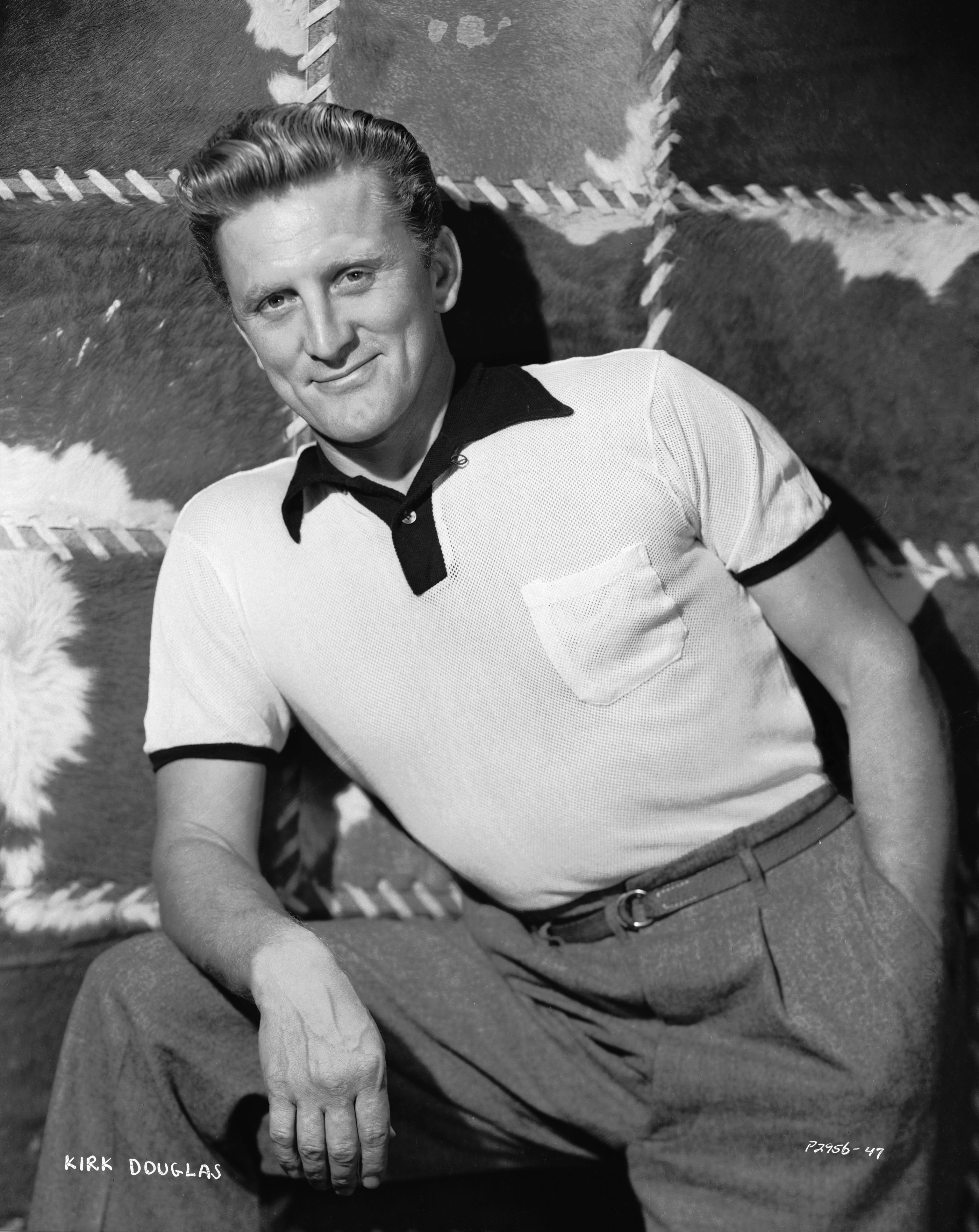 Kirk Douglas posant avec une main dans sa poche tout en posant son autre bras sur ses genoux, portant un t-shirt noir et blanc et un pantalon gris en 1950. / Source : Getty Images