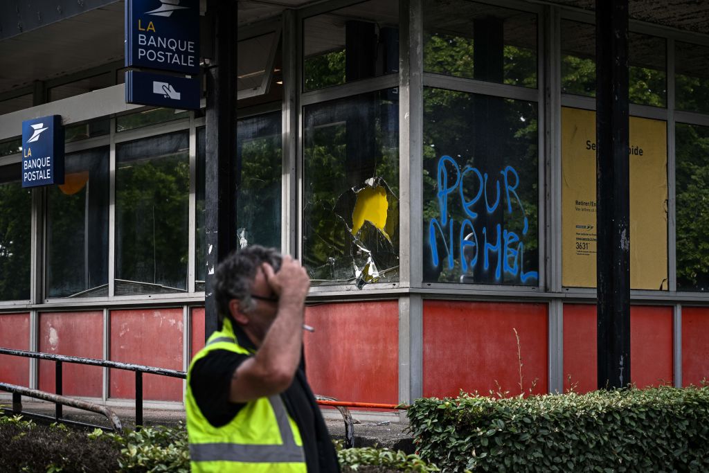 Un ouvrier fume une cigarette à côté d'un bureau de poste endommagé portant un message "Pour Nahel dans le quartier des Aubiers à Bordeaux, dans le sud-ouest de la France, le 30 juin 2023, après les émeutes et les incidents survenus dans tout le pays à la suite du tir à bout portant d'un jeune de 17 ans dans la poitrine par la police à Nanterre, dans la banlieue ouest de Paris.  (Photo by Philippe LOPEZ / AFP) (Photo by PHILIPPE LOPEZ/AFP via Getty Images)