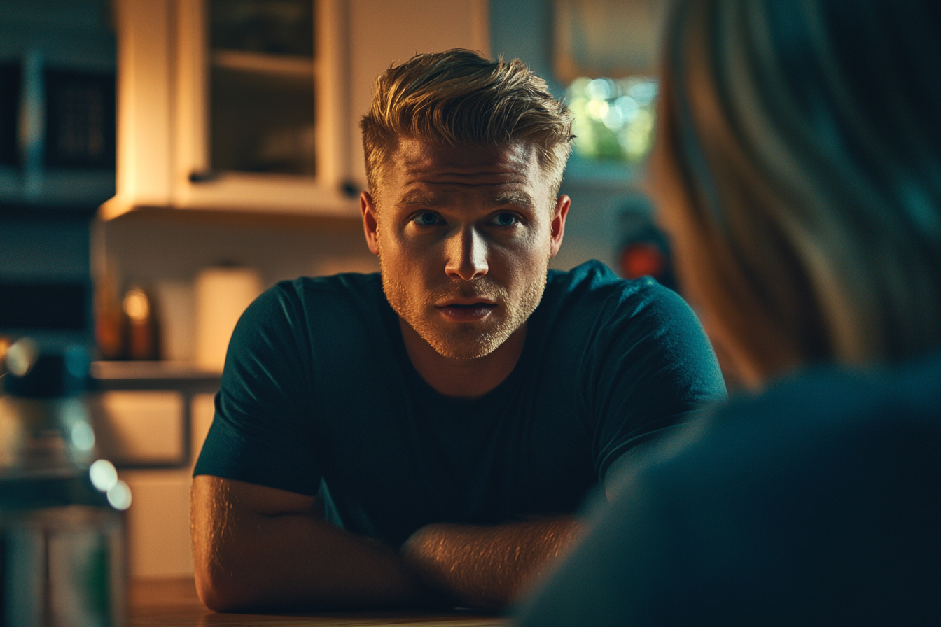 Un homme blond d'une trentaine d'années a l'air inquiet tout en parlant à une femme à la table de la cuisine | Source : Midjourney