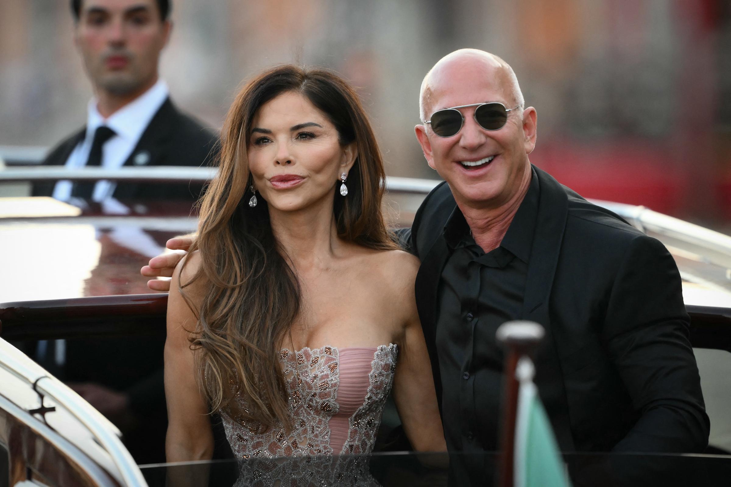 Jeff Bezos affiche un large sourire alors qu'il se penche vers Lauren Sánchez, qui regarde devant elle avec un air serein. Le couple se tient sur le pont du bateau, rayonnant de confiance, tandis qu'un agent de sécurité en costume les observe depuis l'arrière, avec en arrière-plan un paysage urbain flou.