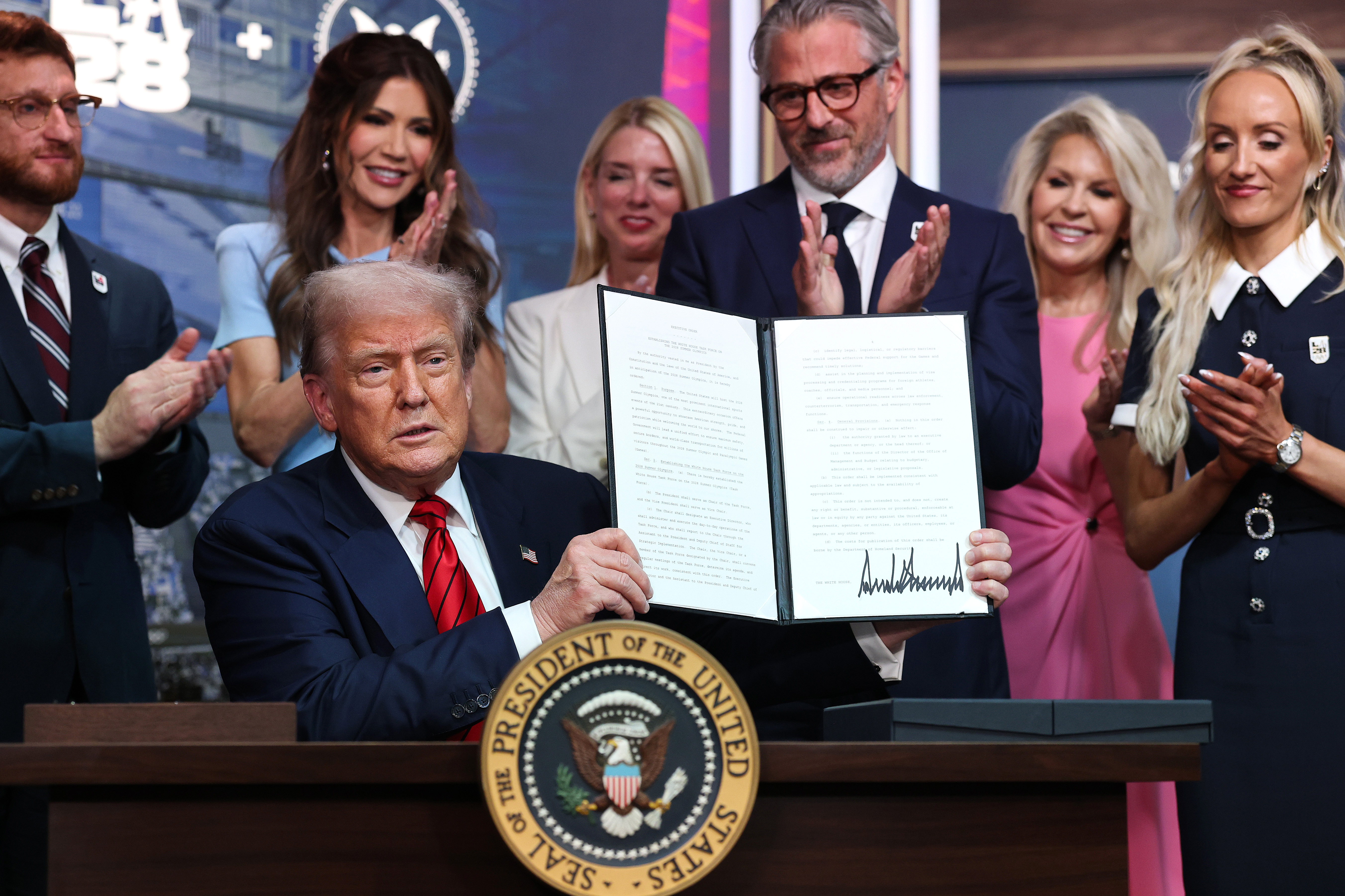 Tenant bien haut le décret signé, Trump le présente au public de l'auditorium South Court de la Maison Blanche, tandis que les fonctionnaires et les invités l'acclament.