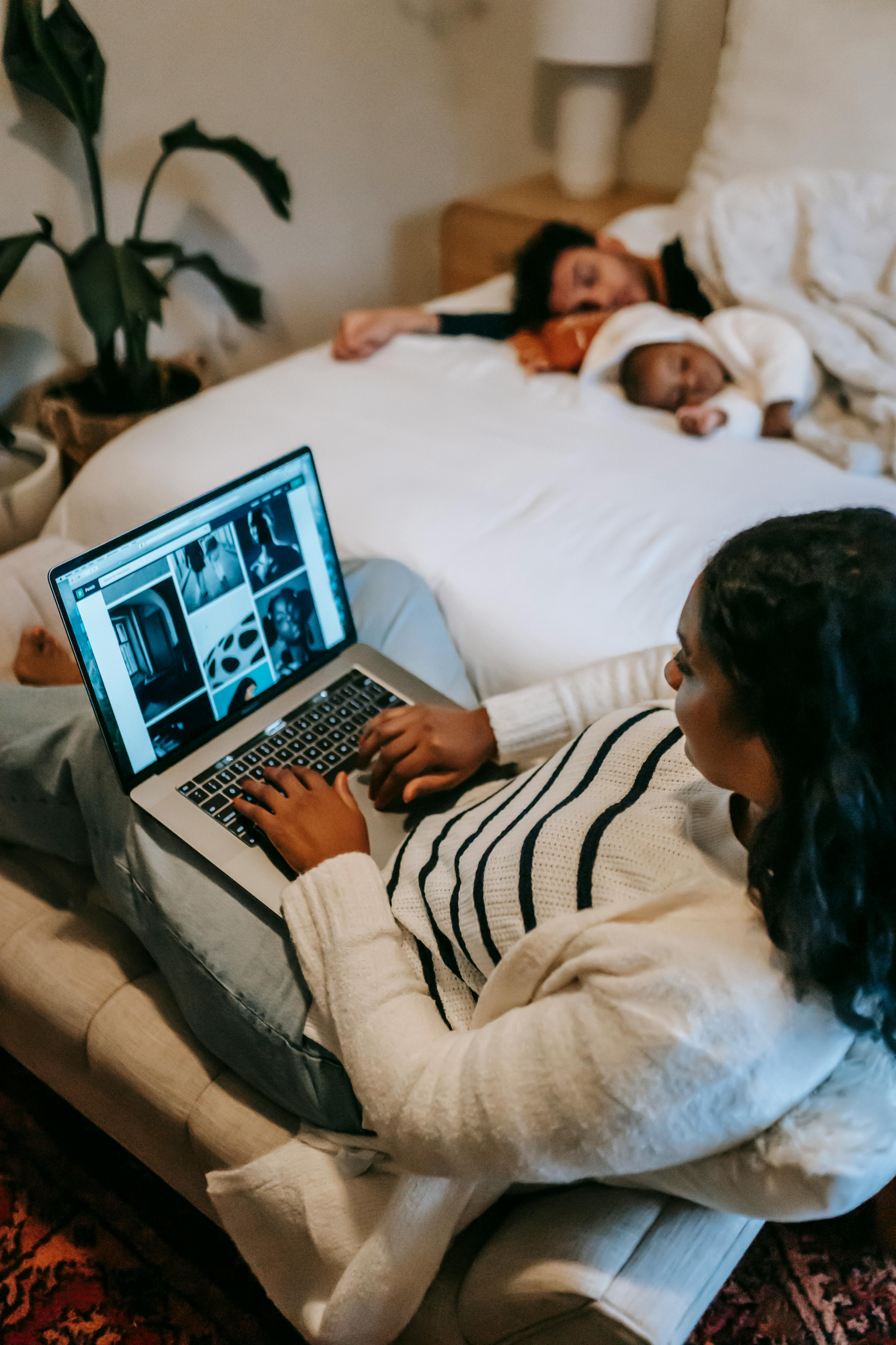 Une femme sur son ordinateur portable et sa famille qui dort sur le lit | Source : Pexels