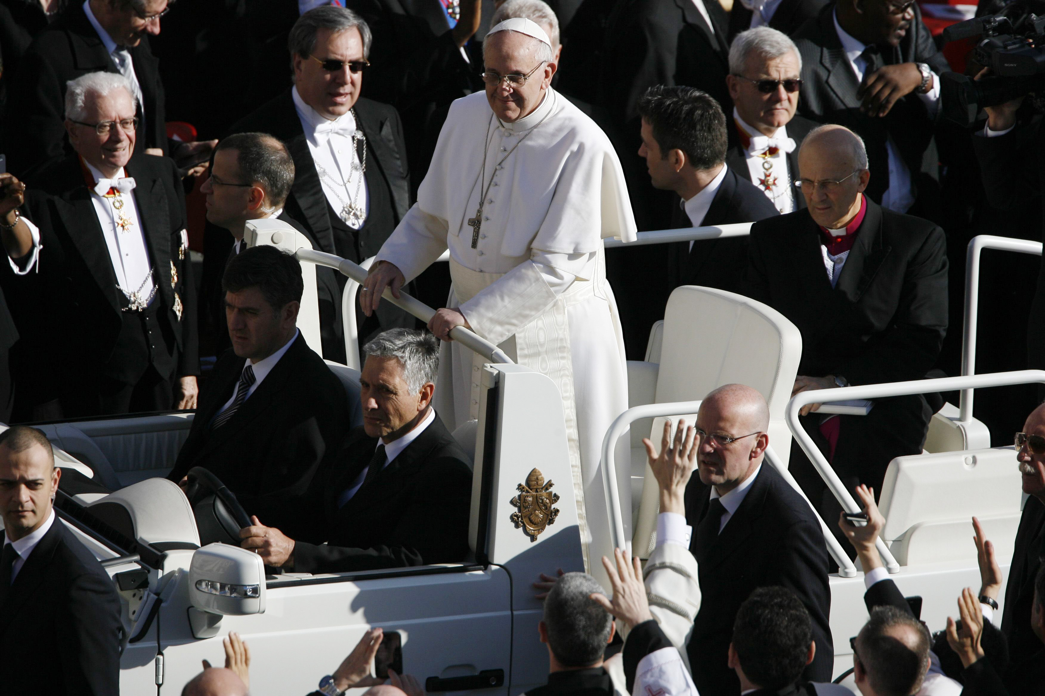 Le pape François se tient dans la papamobile, entouré d'une mer de dignitaires et de mains qui s'agitent, entièrement vêtu de blanc avec une croix argentée drapée sur sa poitrine
