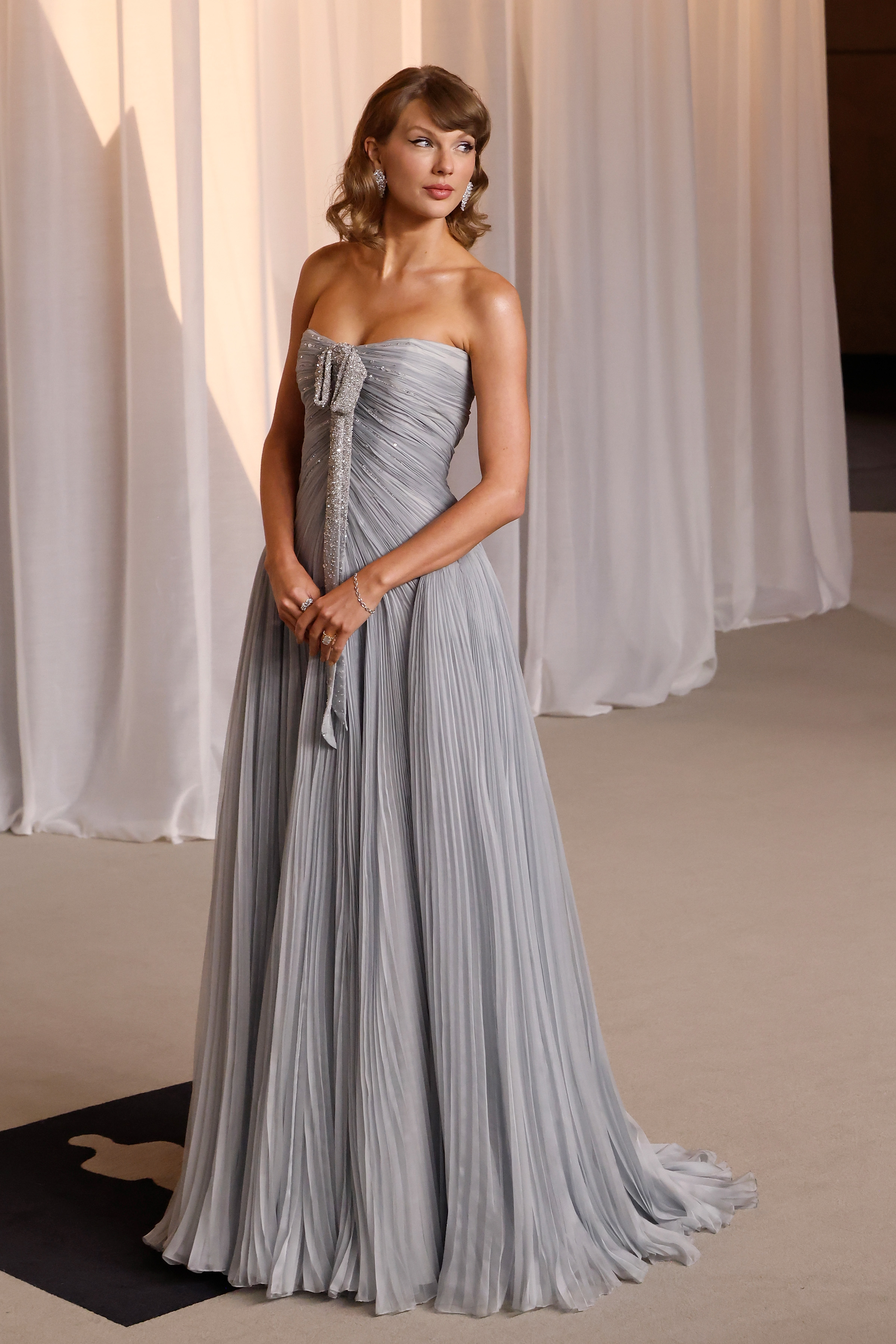 Taylor Swift respire l'élégance éthérée dans une robe bustier gris tourterelle, rehaussée de plis délicats sur tout le corsage et la jupe ample. Un nœud scintillant, orné de cristaux, descend gracieusement sur le devant, rehaussé par des boucles d’oreilles pendantes en diamants étincelantes et une chevelure ondulée au look vintage tout en douceur. Prête à monter sur scène en tant que tête d’affiche de la soirée, la lauréate d’un Grammy Award incarne un glamour intemporel à l’état pur.