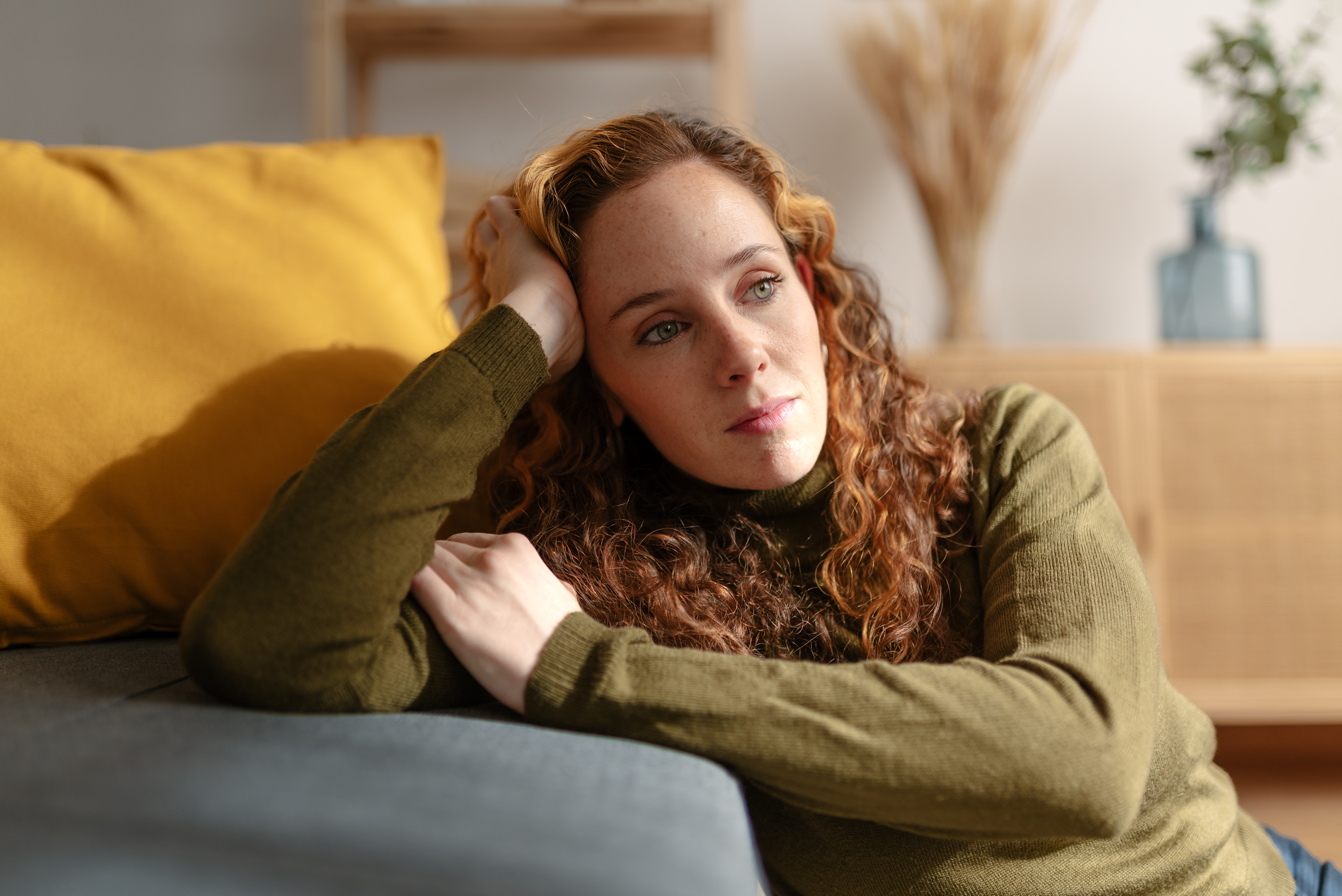 Portrait d'une jeune femme aux longs cheveux roux bouclés, assise par terre et appuyée sur un canapé, regardant au loin, le visage triste. | Source : Getty Images
