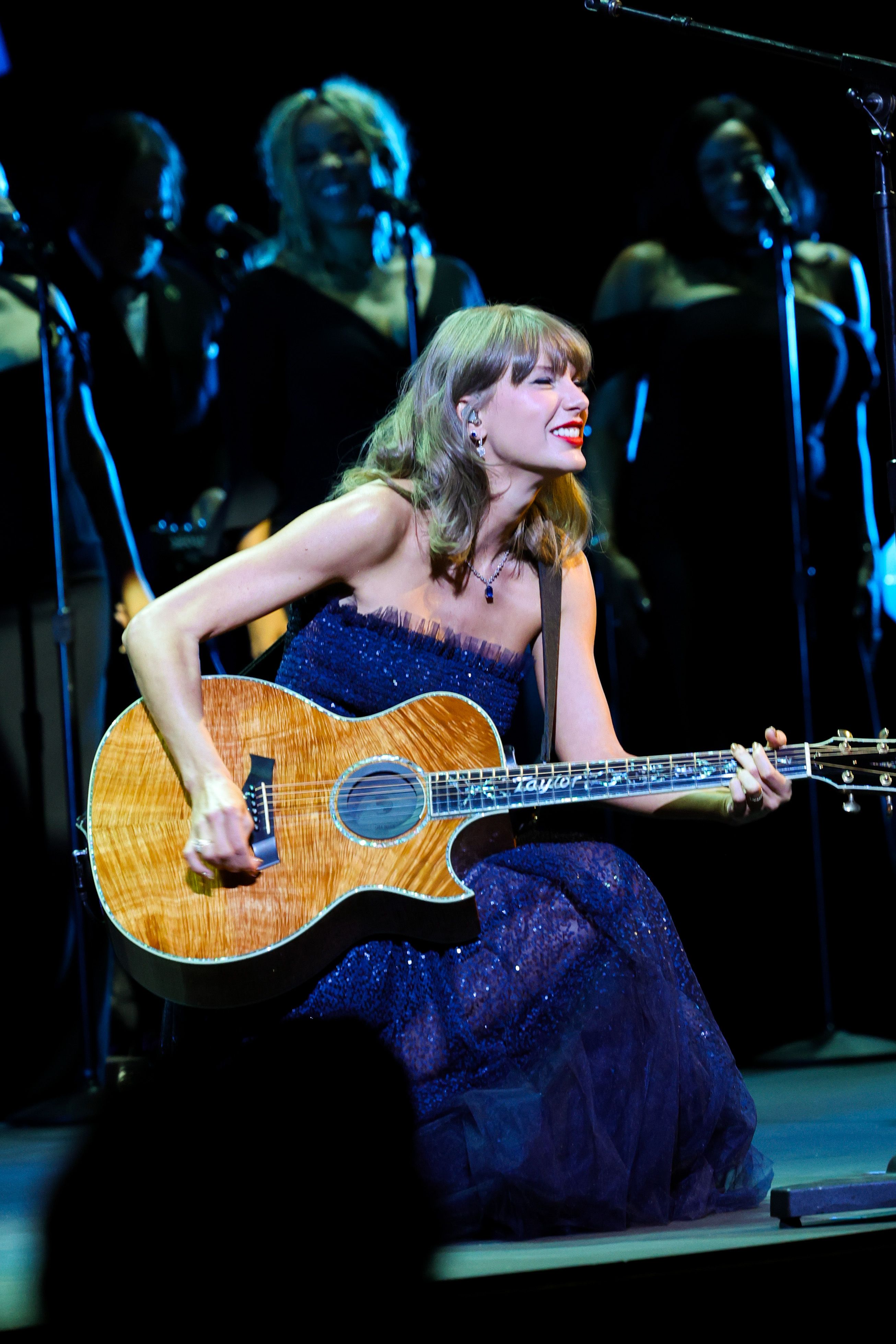 Taylor Swift donne un concert acoustique dans une robe bustier bleu nuit, scintillante de paillettes sur toute la surface et ornée d'une délicate bordure en tulle au niveau du décolleté. Assise avec sa guitare acoustique sur mesure, qui est sa marque de fabrique, la chanteuse associe cette tenue de scène étincelante à un délicat collier pendentif en saphir, des boucles d'oreilles pendantes en diamant, son rouge à lèvres rouge classique et de douces ondulations blond miel.