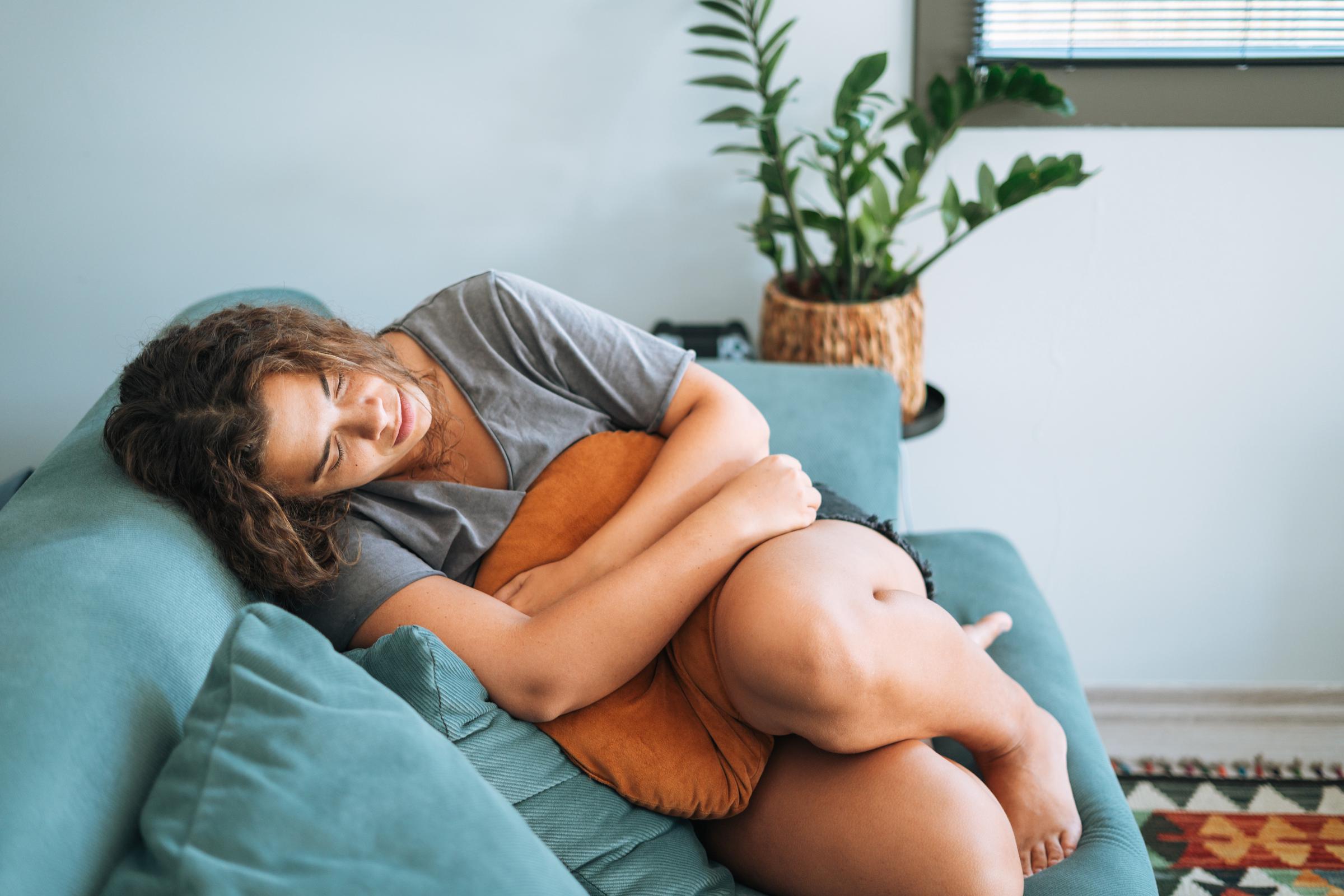 Une jeune femme est allongée sur un canapé, tenant un oreiller contre son abdomen, semblant ressentir de la douleur | Source : Getty Images