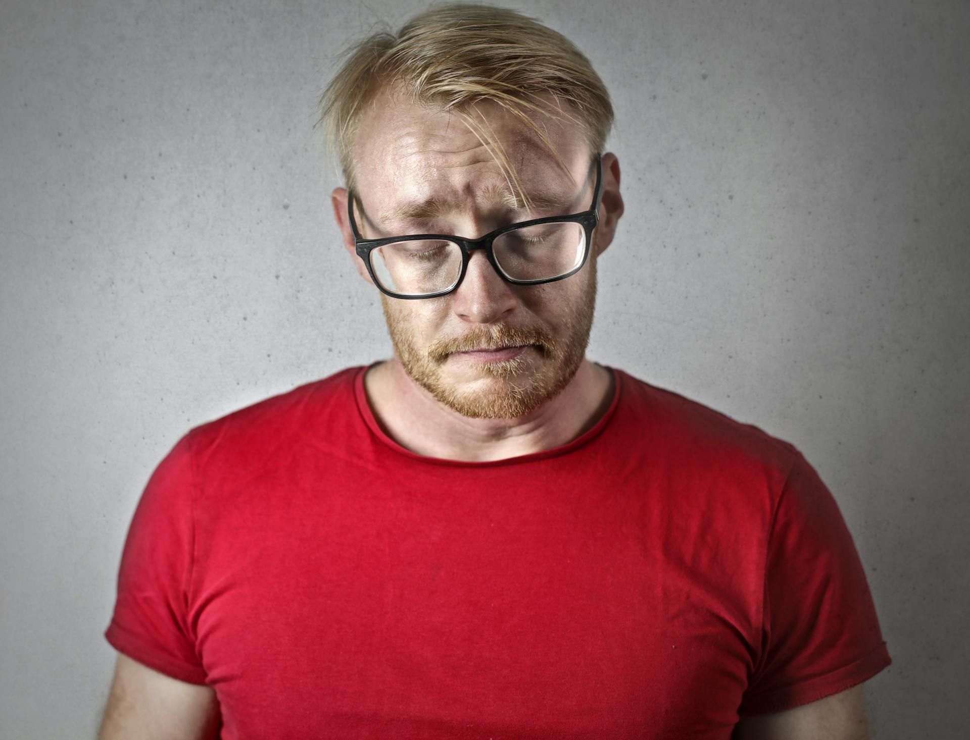 Un homme triste en T-shirt rouge et lunettes à monture noire fermant les yeux | Source : Pexels