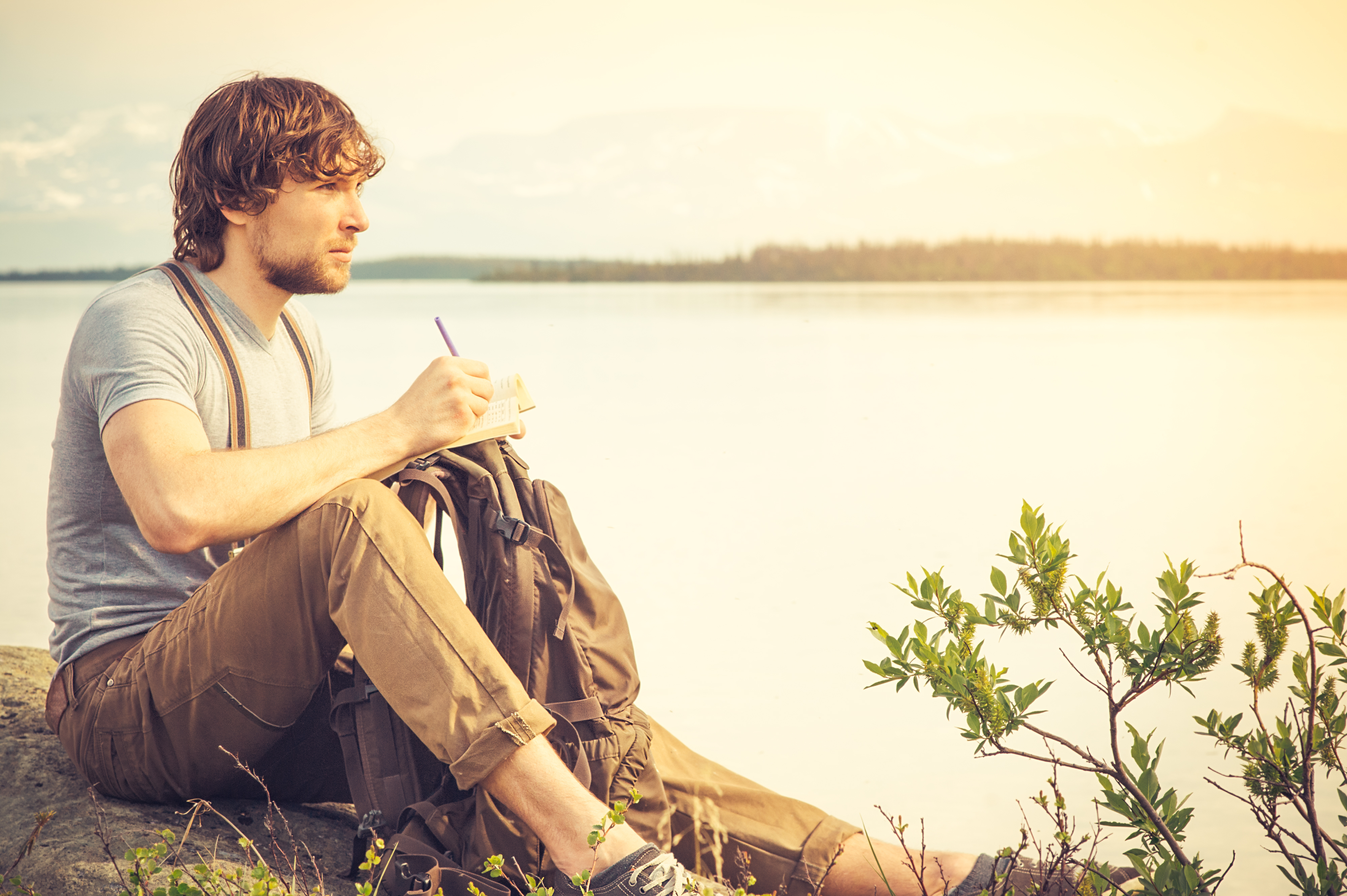 Jeune homme lisant un livre en plein air avec un lac en arrière-plan | Source : Getty Images