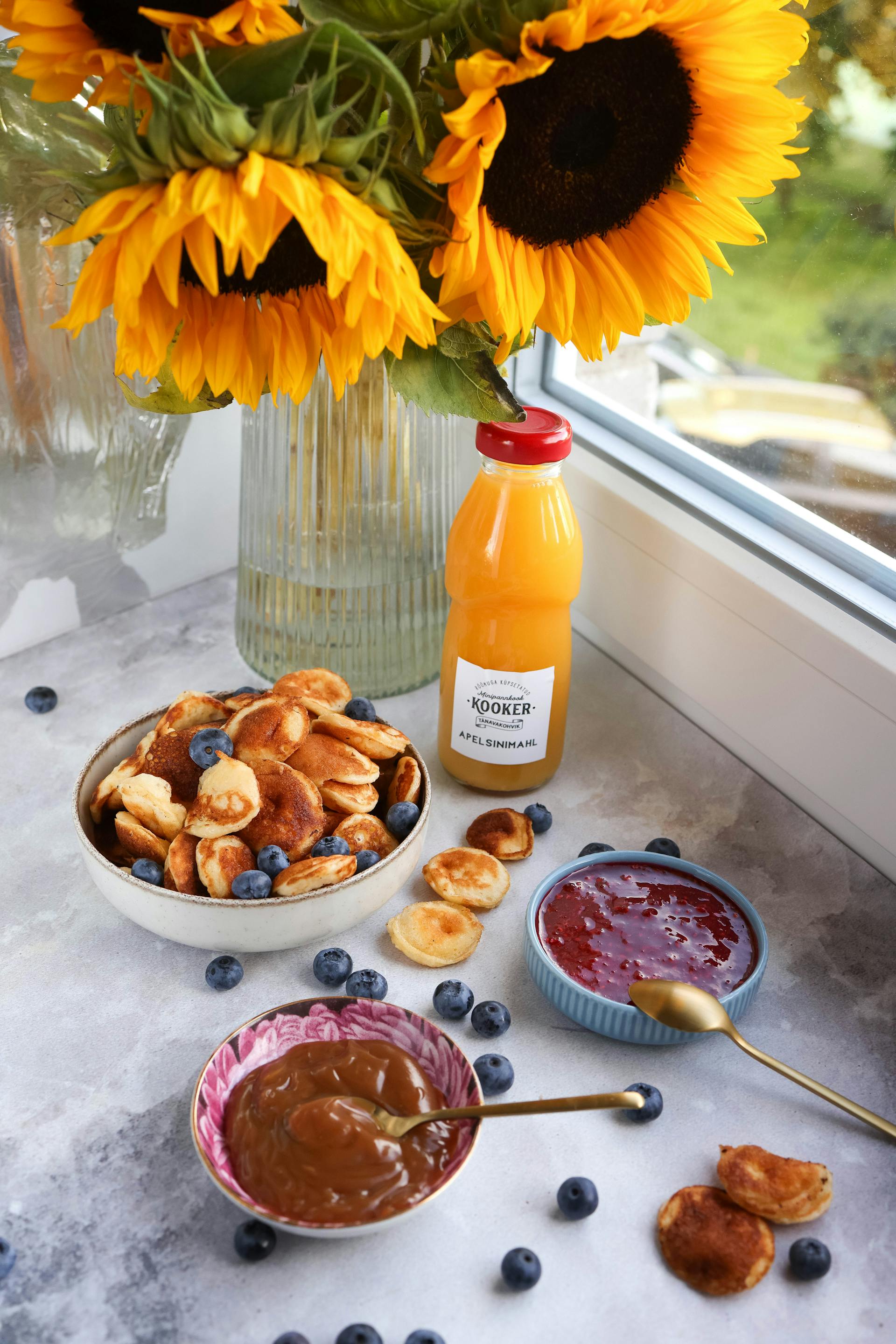 Une photo montrant des crêpes avec des myrtilles et du jus de fruits reposant près d'un vase de tournesols à côté de la fenêtre de la cuisine | Source : Pexels