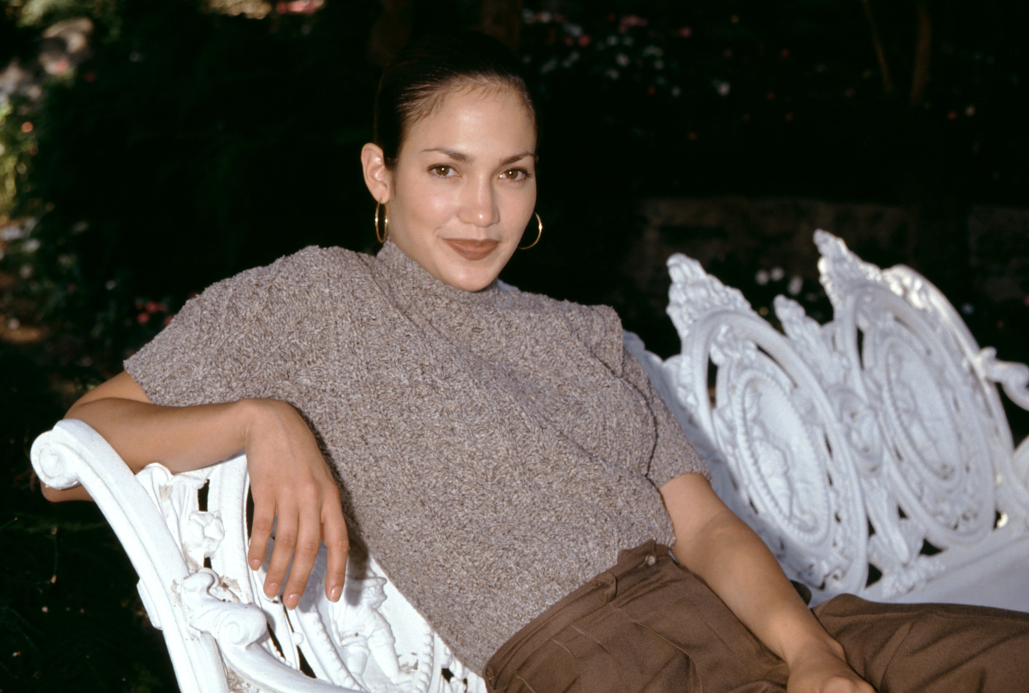 Jennifer Lopez est allongée nonchalamment sur un banc blanc orné, reposant un bras le long du cadre sculpté, vêtue d'un haut texturé à encolure haute et d'un pantalon marron ample. Avec ses cheveux gominés, ses boucles d'oreilles et son expression calme, le portrait capture un moment décontracté mais poli de début de carrière.